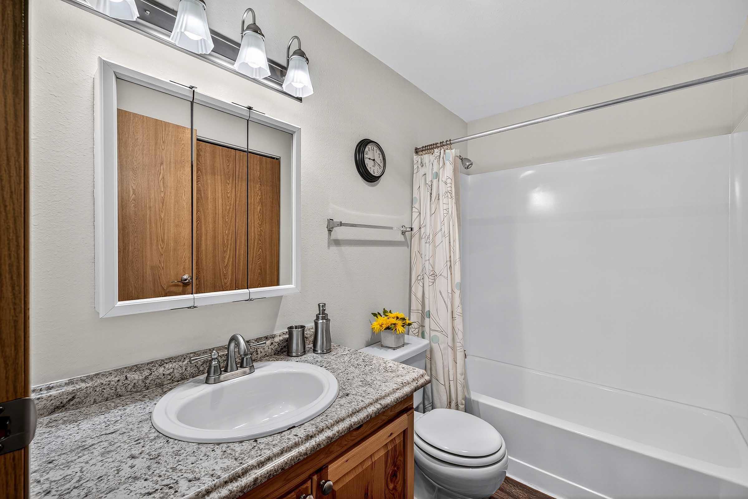 A clean bathroom featuring a white bathtub, a granite countertop sink with modern fixtures, a mirror, and a clock on the wall. There are towels hanging on a bar, a shower curtain with a floral design, and a small vase with yellow flowers on the counter. Soft, neutral-colored walls create a calm atmosphere.