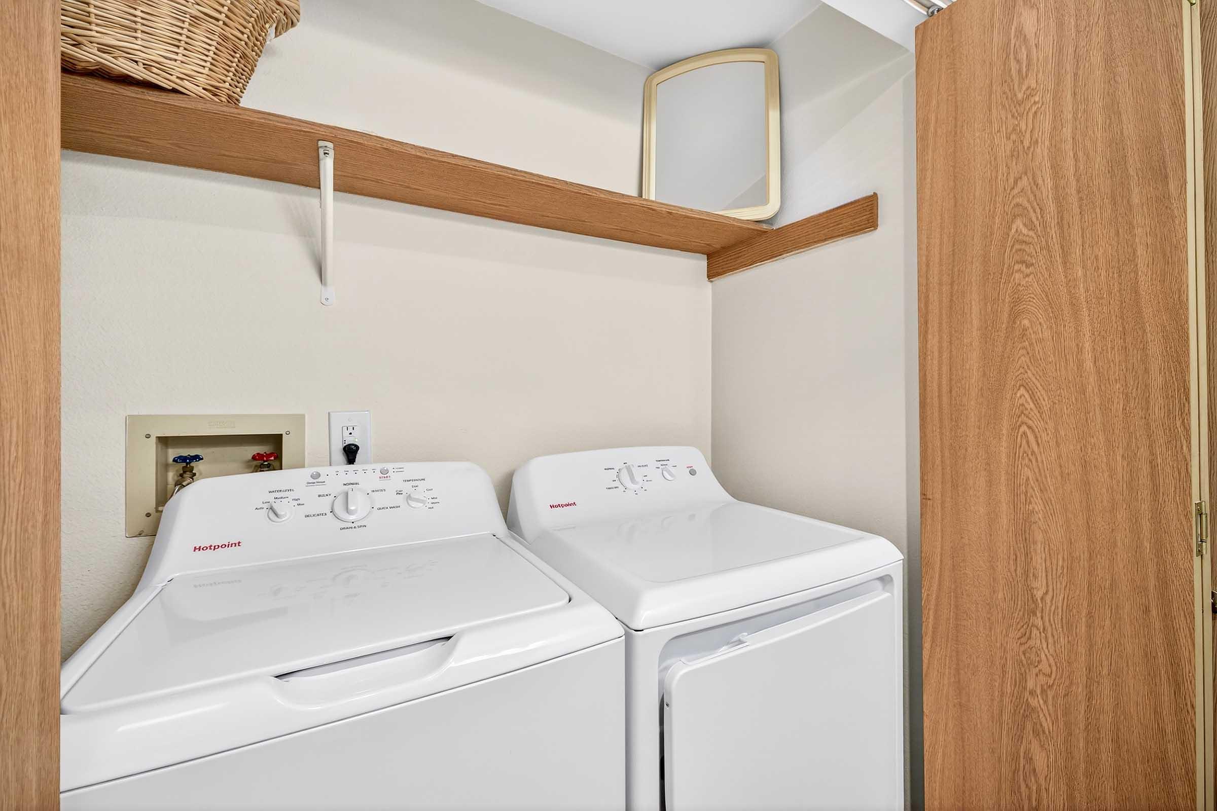 A laundry area featuring stacked white washing machine and dryer units, a wooden shelf above, and a wicker basket on the shelf. The wall is a light color, and there is a small mirror above the shelf. The space is compact and organized.
