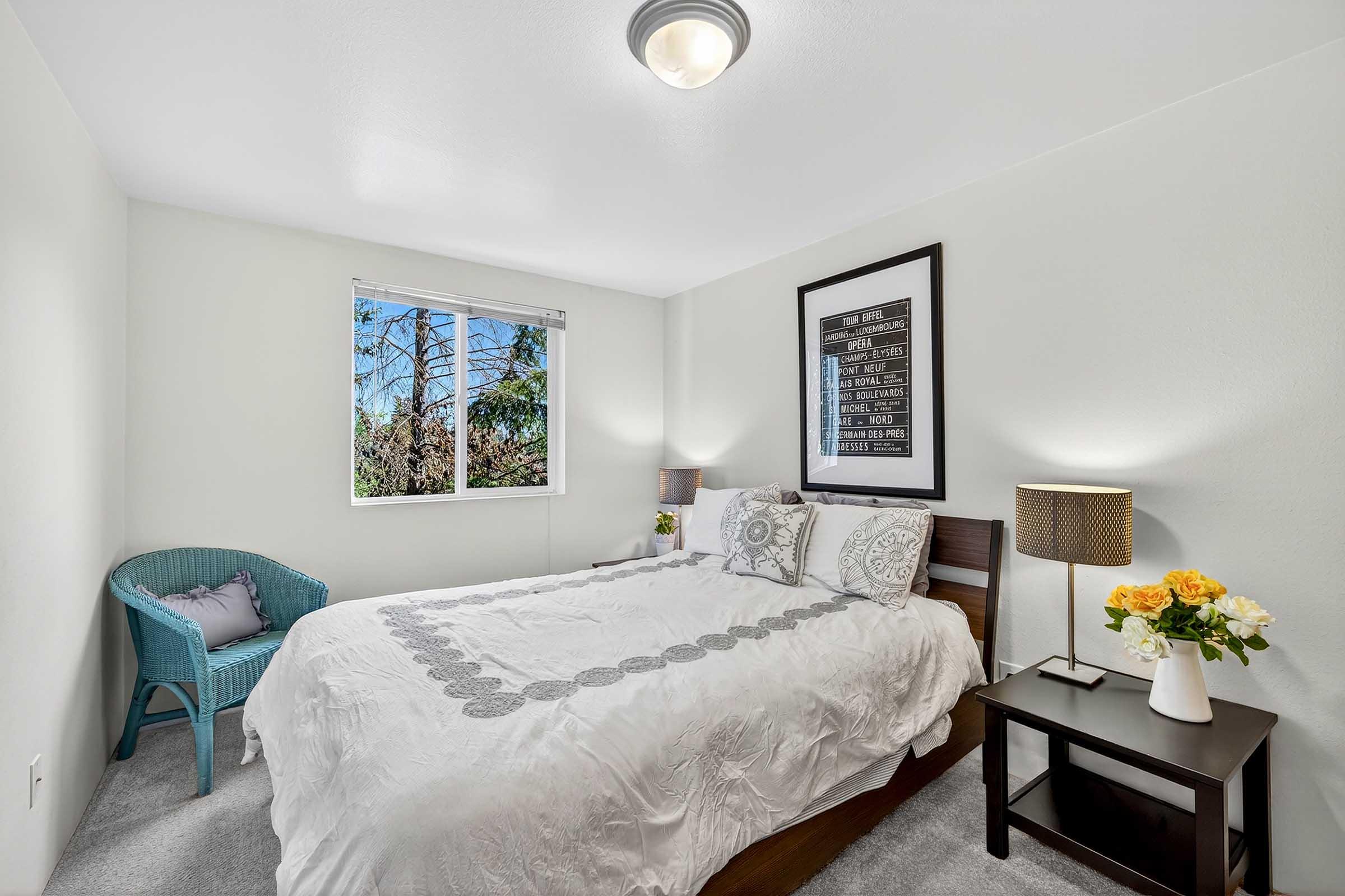 Cozy bedroom featuring a double bed with white bedding, a bedside table with a lamp and a vase of flowers, a small teal chair, and a window allowing natural light in. The walls are painted white, creating a bright and airy atmosphere.