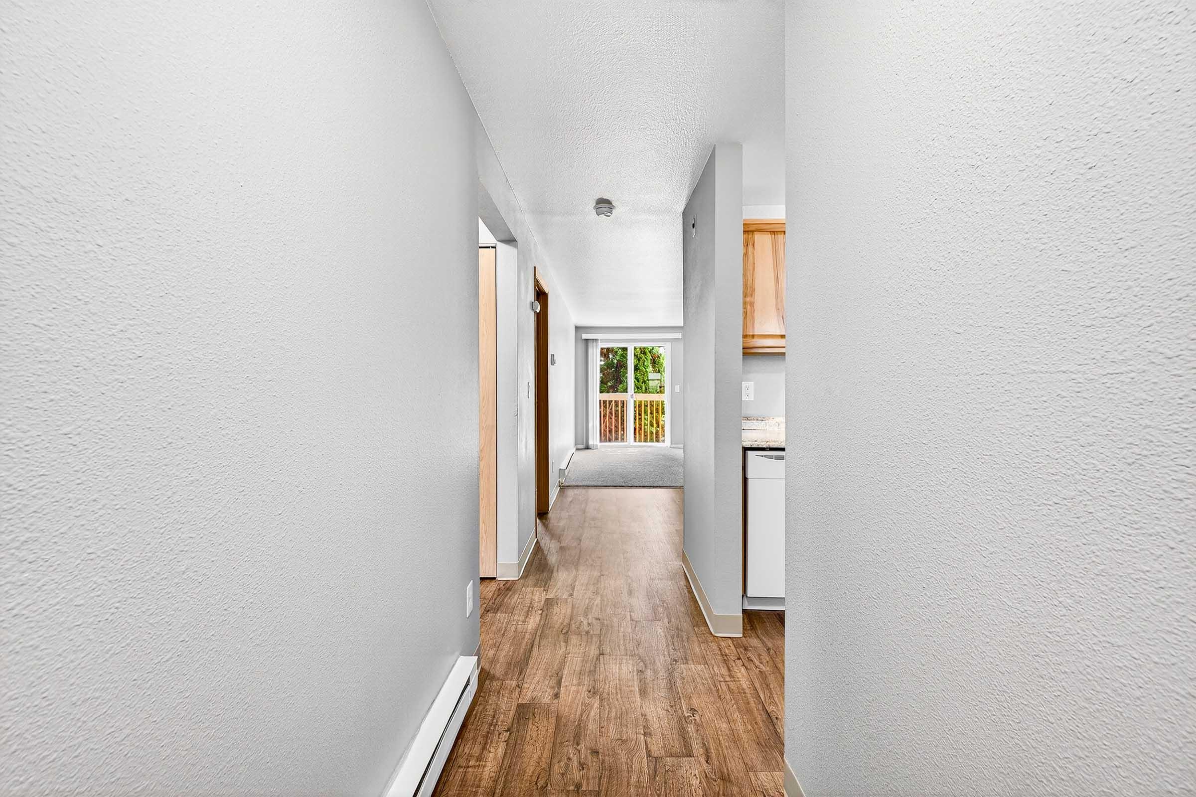 A spacious hallway in a light-colored apartment with gray walls and hardwood flooring. At the end of the hallway, there is a view of a sliding glass door leading to a balcony. The kitchen area to the right features wooden cabinets, and the overall ambiance is bright and inviting.