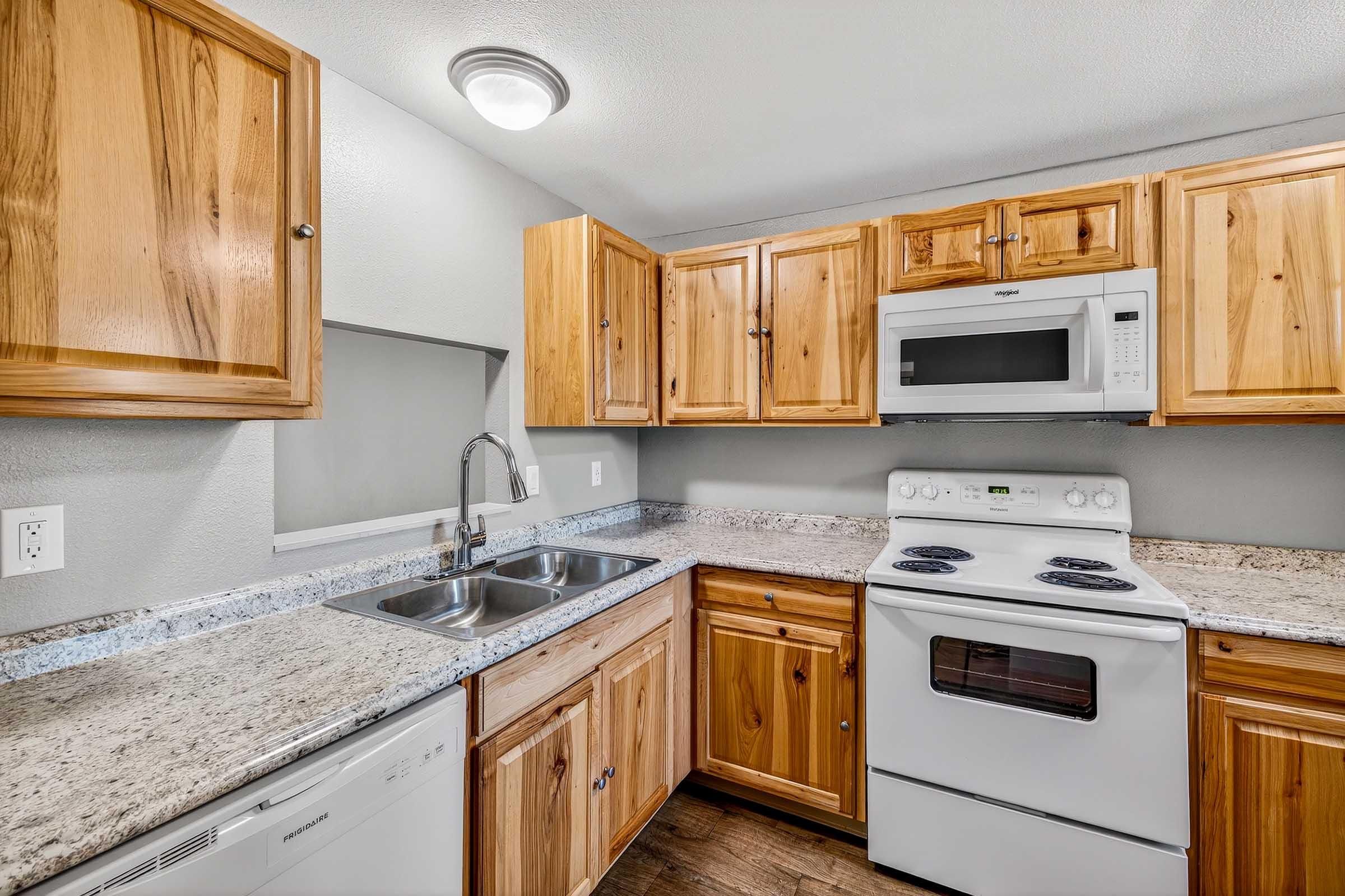 A modern kitchen with wooden cabinets, a double sink, and granite countertops. It features a white stove, microwave, and dishwasher, along with overhead lighting. The walls are painted gray, and the overall aesthetic is bright and inviting.