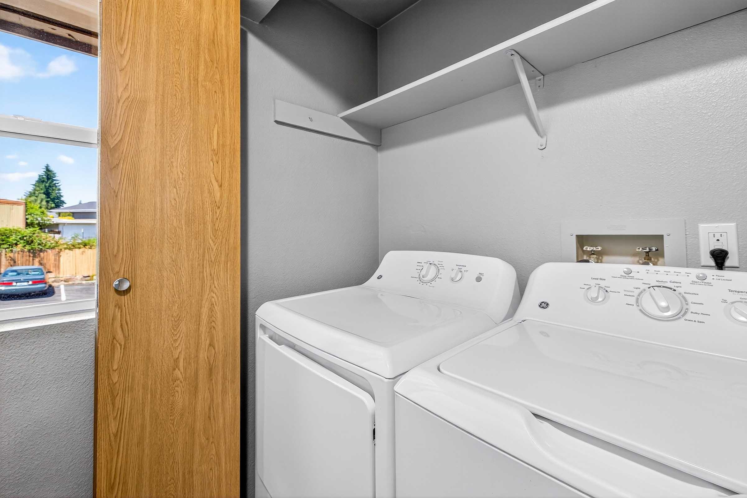 A laundry room featuring a washing machine and dryer side by side. There is a window with a view of the outside, a wooden door, and a shelf above the appliances. The walls are painted in a light color, creating a clean and organized space.