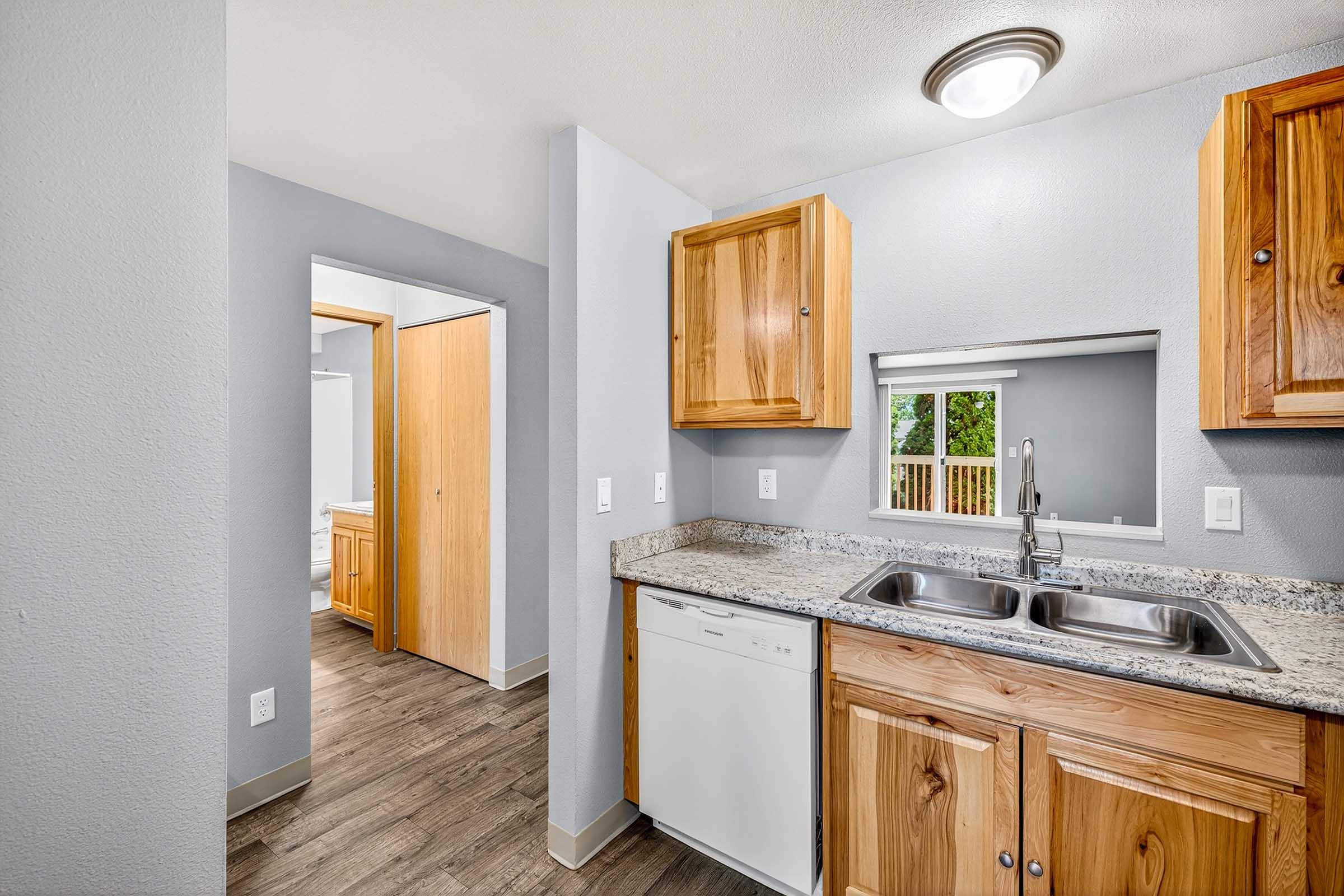Modern kitchen with wooden cabinets, a double sink, and a dishwasher. The space features a granite countertop and an open design leading to a bathroom. Light walls and laminate flooring create a bright and inviting atmosphere. Natural light enters through a nearby window.