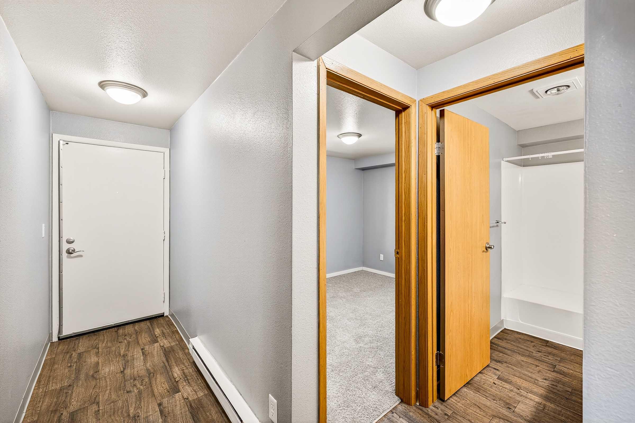 A hallway with two doorways, one leading to an empty room and the other to an empty bathroom. The walls are painted light gray, and the flooring is a dark wood laminate. There are flush-mounted ceiling lights, and the doors are wooden with a light finish. The space appears well-lit and clean.