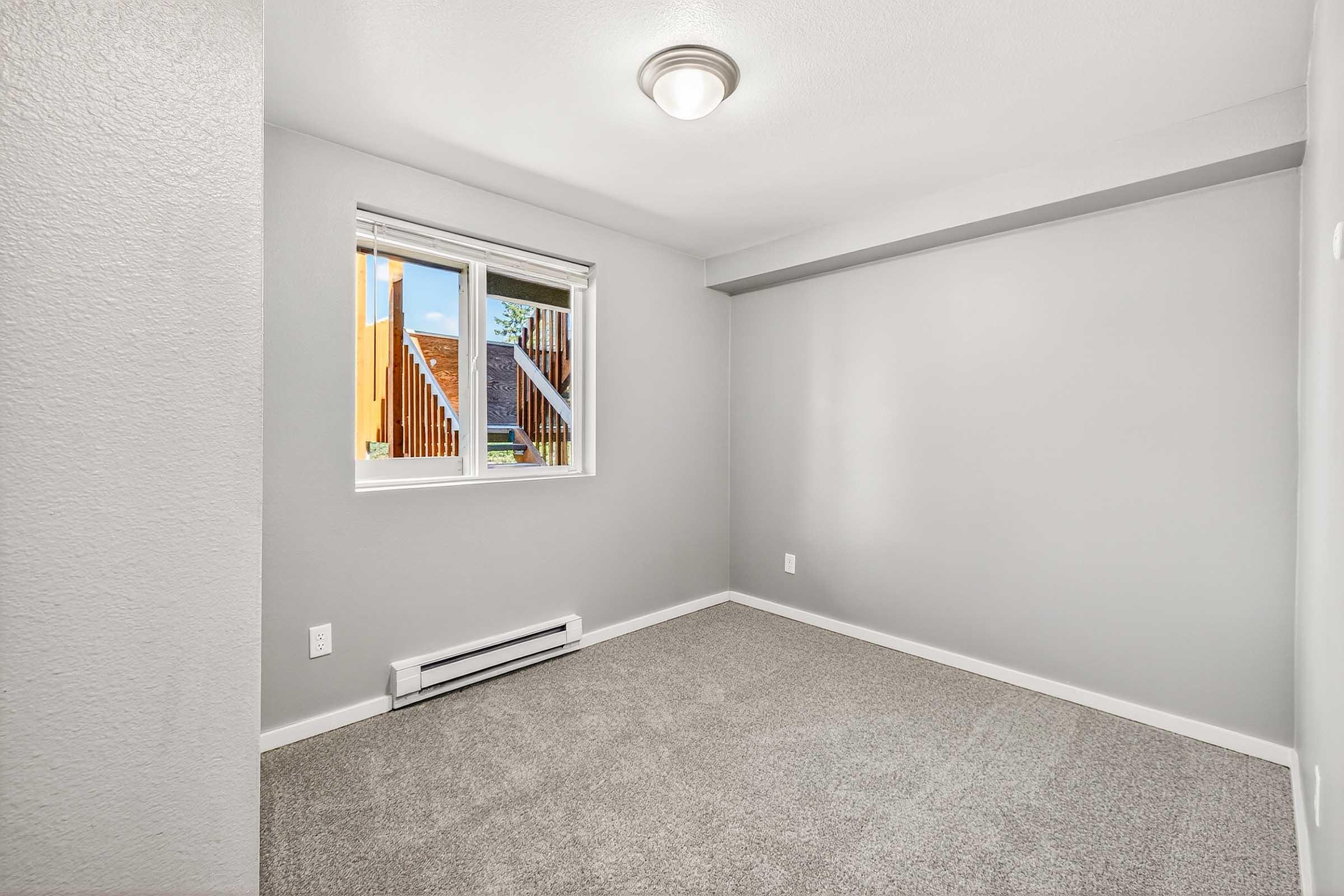 Empty room with light gray walls and carpet. A window with white blinds allows natural light in, showing wooden exterior elements outside. There is a ceiling light fixture and baseboard heating visible. The overall atmosphere is clean and minimalistic.
