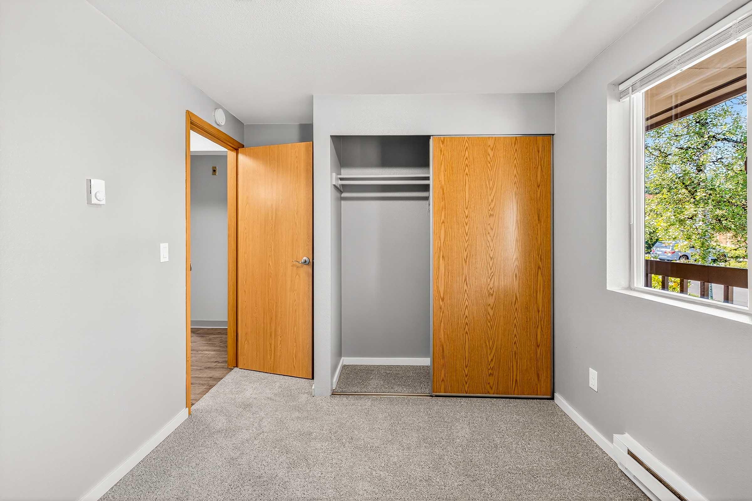 A small, well-lit room featuring light gray walls and carpet. The room includes a wooden door open to a closet with a hanging rod and a window with a view of trees outside. A light switch is visible on the wall near the entrance.