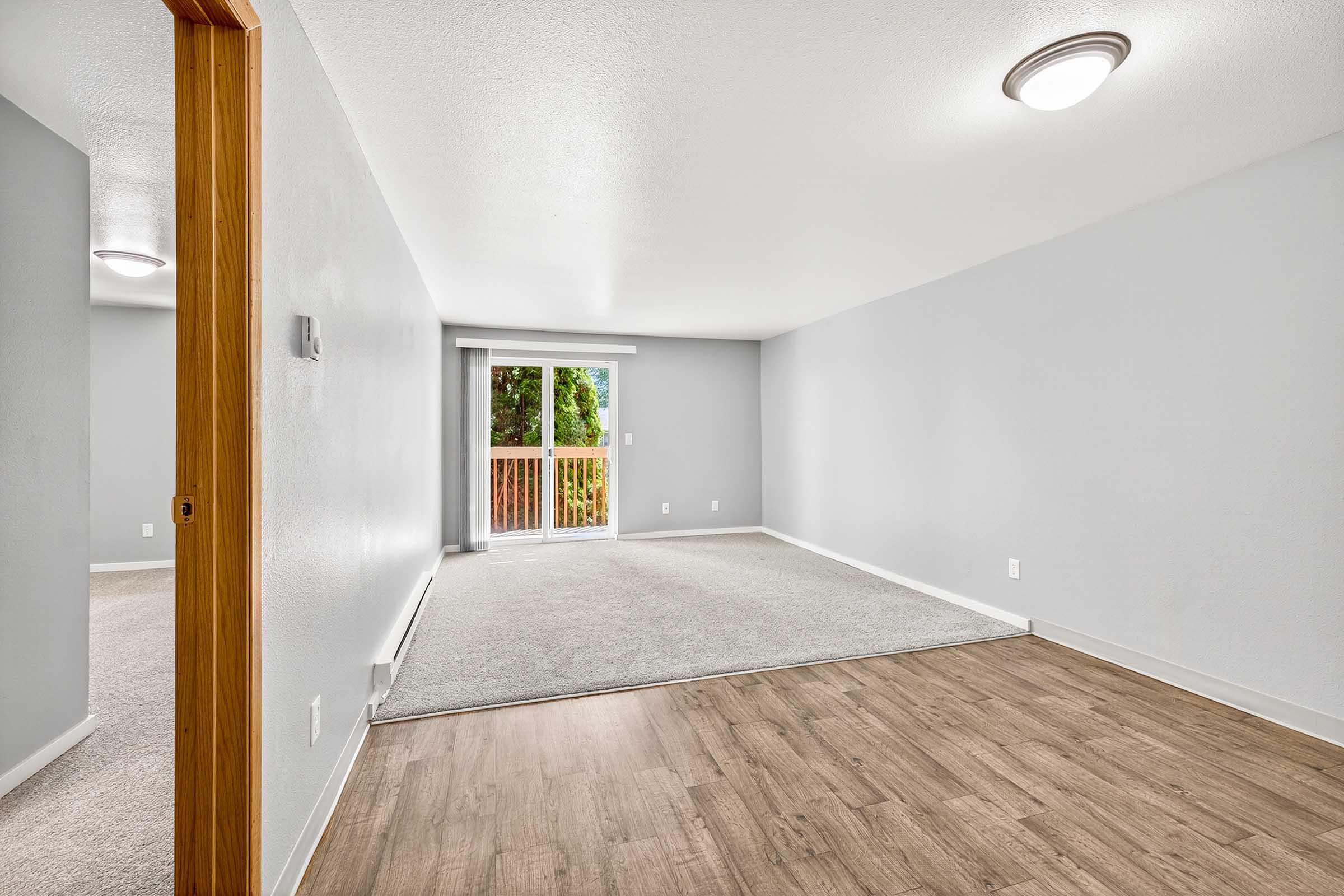 A spacious interior view of a light gray room with carpeted and laminate flooring. A balcony is visible in the background, featuring sliding glass doors. The walls are painted light gray, and there are modern ceiling lights. The overall ambiance is bright and inviting, ideal for a living space.