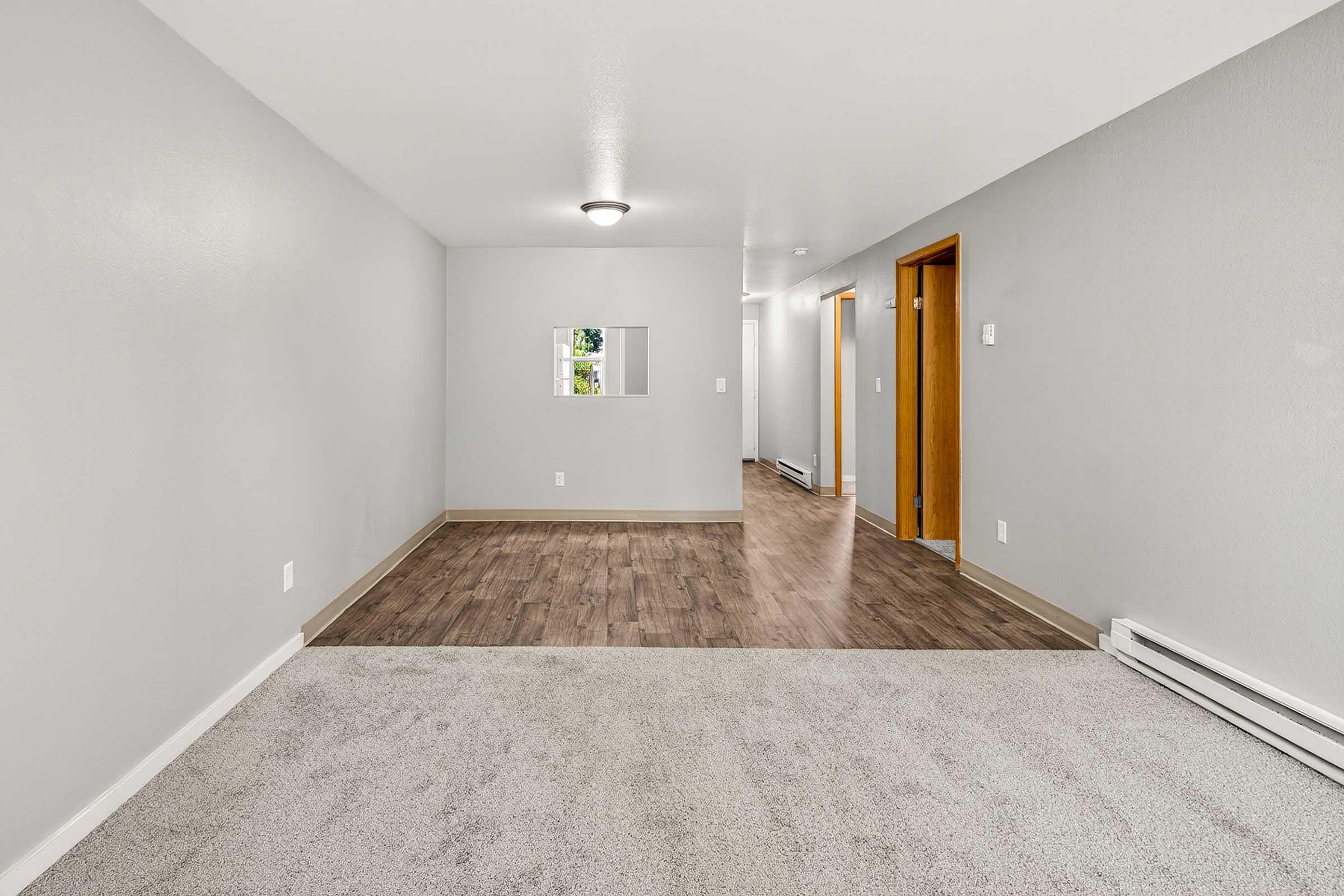 A spacious, light-filled room with gray walls and carpeted flooring. The area features a small window in the wall, two doorways leading to other rooms, and a well-defined layout. The flooring transitions from carpet to wood, creating an inviting atmosphere.