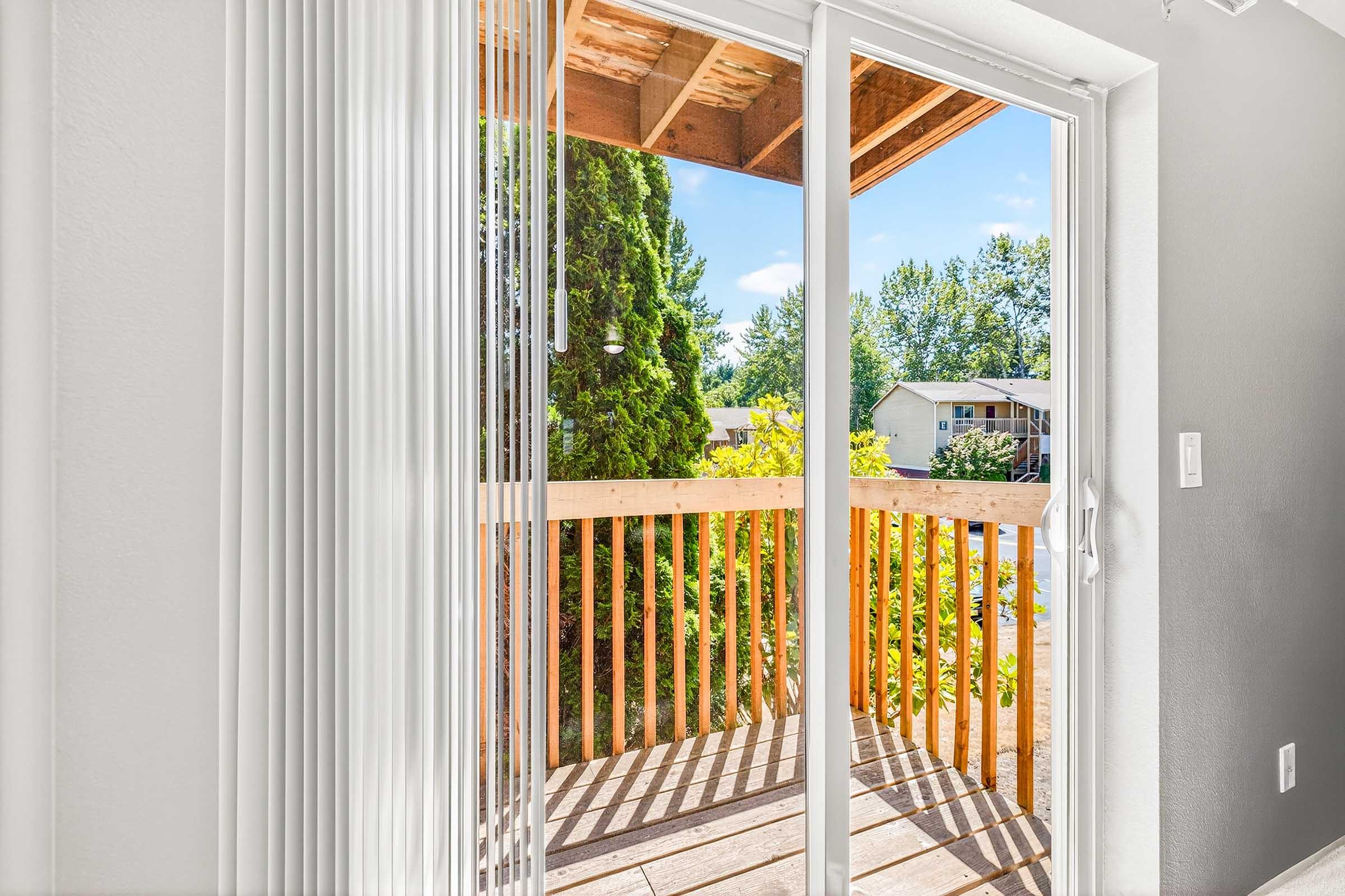 View from an interior space showcasing sliding glass doors that open to a small wooden balcony. The balcony overlooks lush greenery and colorful trees, with blue skies in the background. Soft light illuminates the area, highlighting the clean and modern design of the room.