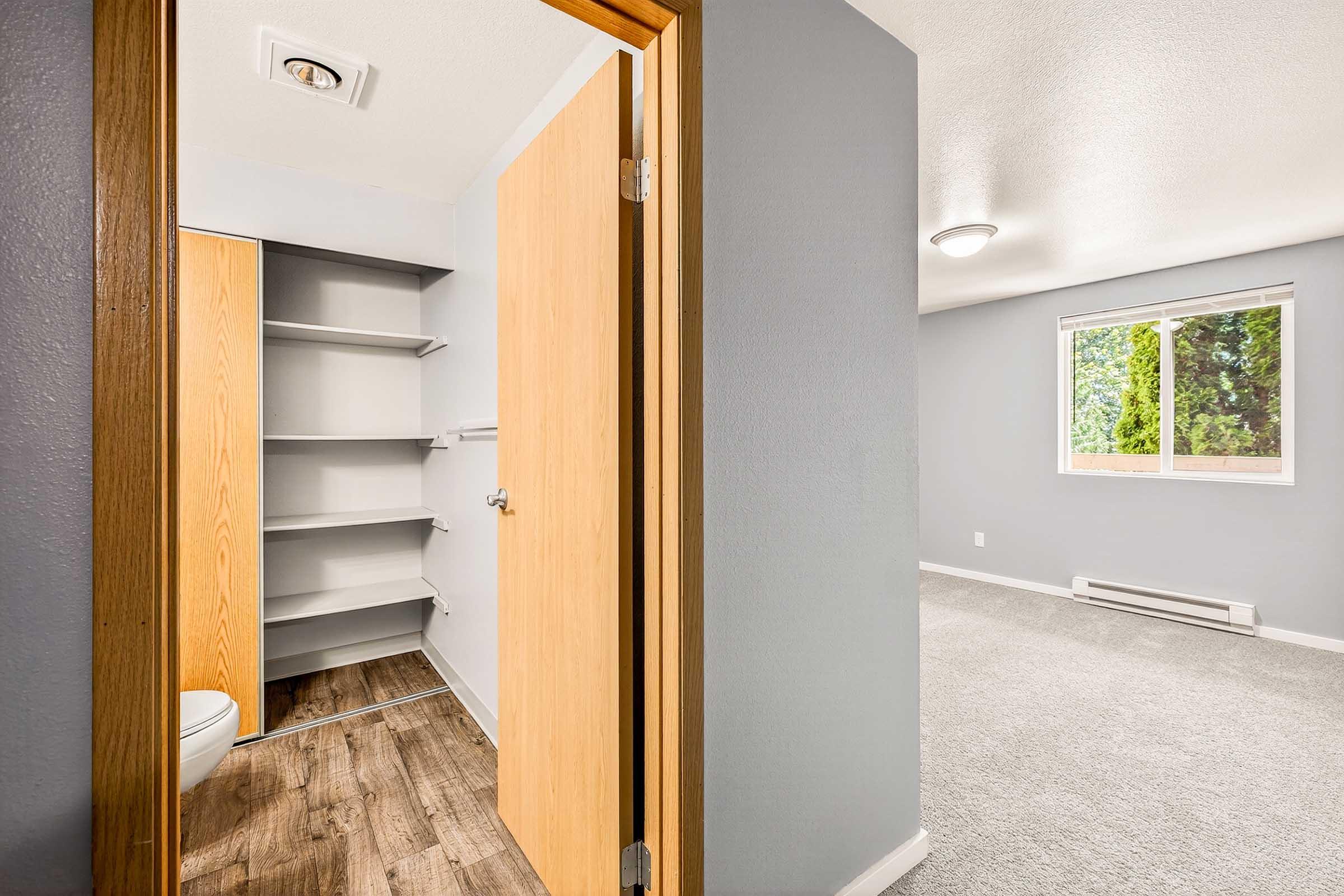 View of a room from a doorway, featuring a partially open door leading to a closet space. The other side shows a carpeted area with a window allowing natural light, painted in soft gray tones. The room appears spacious and well-lit, with neutral-colored walls and laminate flooring.