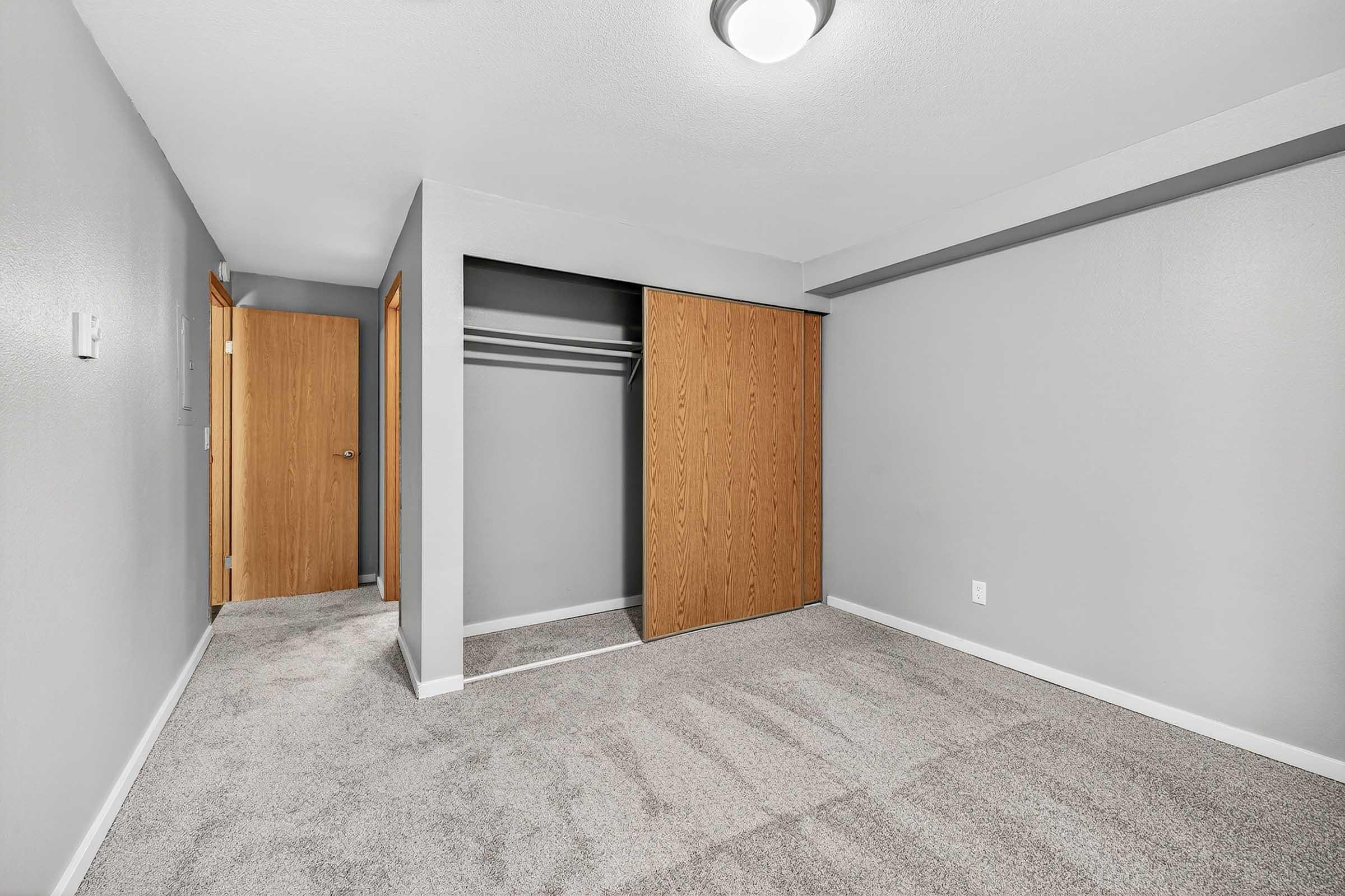 A well-lit, empty bedroom featuring gray walls and carpeted flooring. There is a large closet with sliding wooden doors on one side, and a door leading to another area in the background. The space appears clean and freshly painted, suitable for furniture arrangement.
