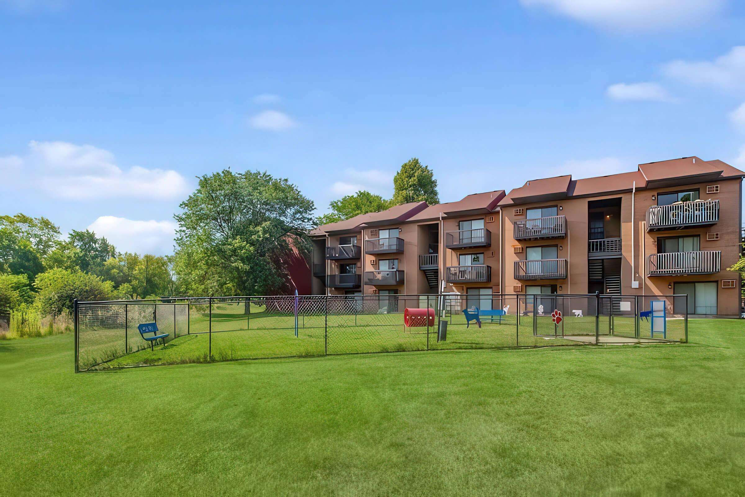 A well-maintained apartment building with multiple balconies overlooks a green yard. There's a fenced area with playground equipment, including swings and a slide, set against a backdrop of trees and a clear blue sky. The scene conveys a vibrant and inviting outdoor space for residents.