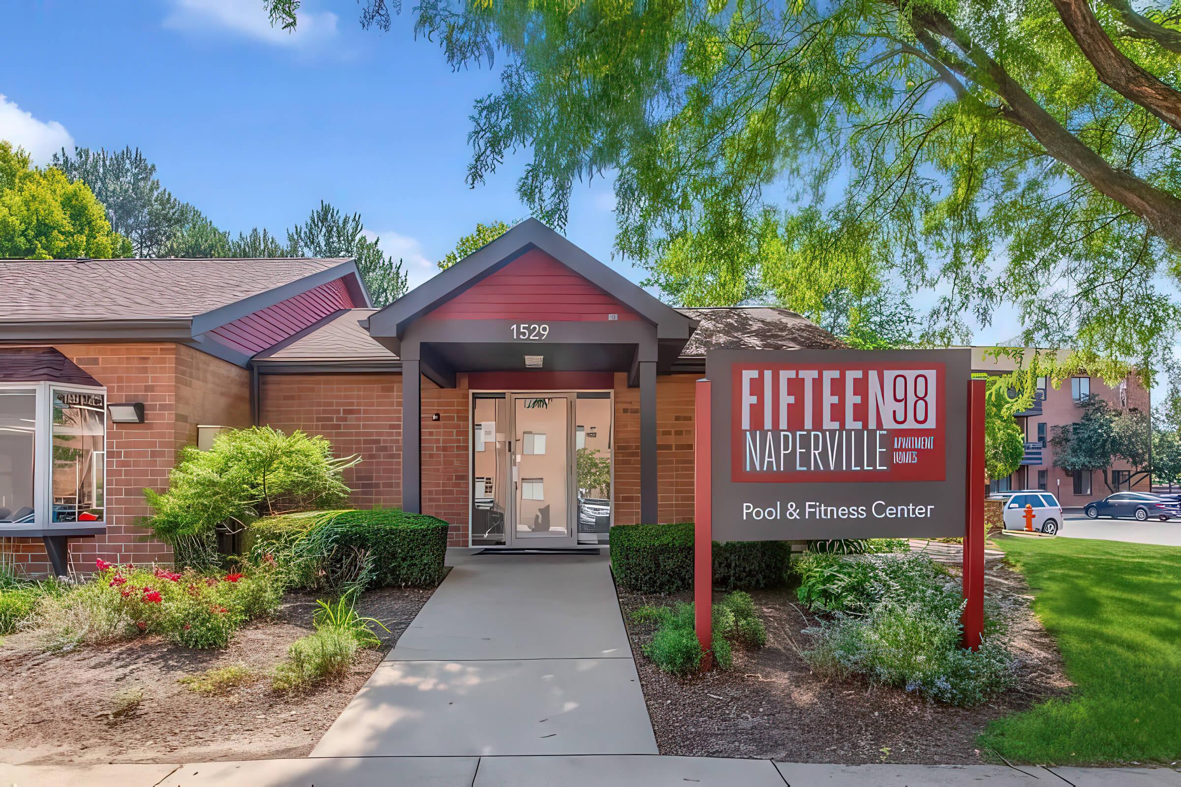 Front view of a building named "Fifteen 98 Naperville," located at 1529, featuring a sign for the Pool & Fitness Center. The structure has a mix of brick and wood accents, surrounded by green landscaping and trees, with a clear blue sky above.