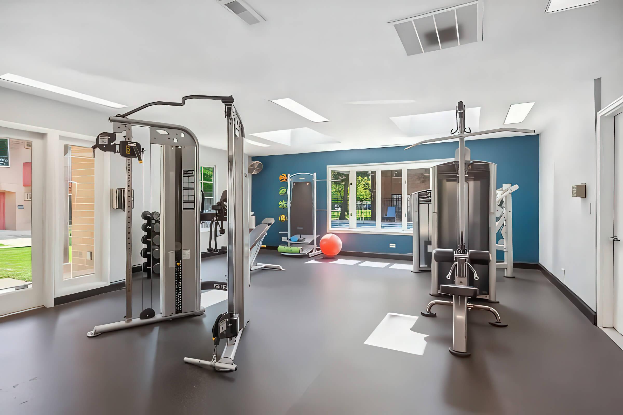 A modern gym interior featuring various exercise equipment including weight machines, a bench, and a fitness ball. Large windows provide natural light, and vibrant wall colors create an inviting atmosphere for workouts. The space is clean and well-equipped for fitness training.