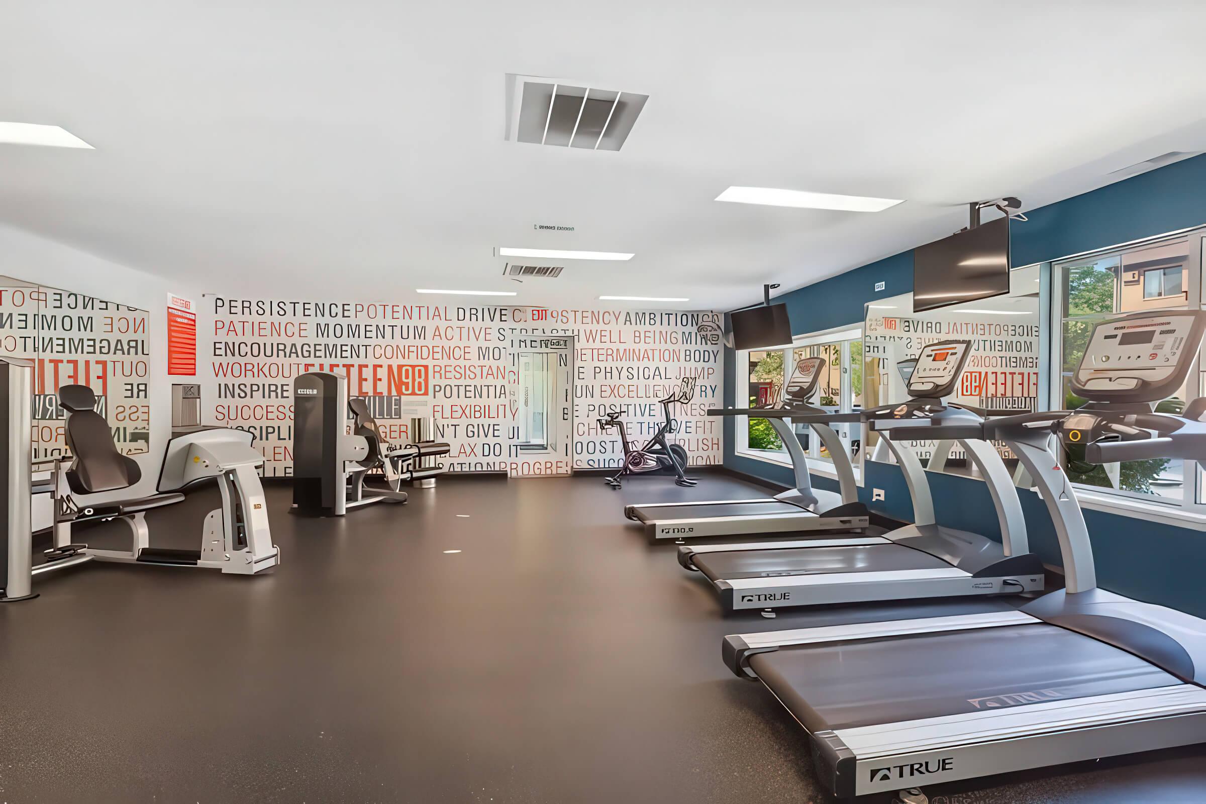 A modern fitness room featuring multiple treadmills, a stationary bike, and a strength training machine. The walls are decorated with motivational words and phrases related to fitness and ambition. Large windows allow natural light, creating an inviting workout environment.