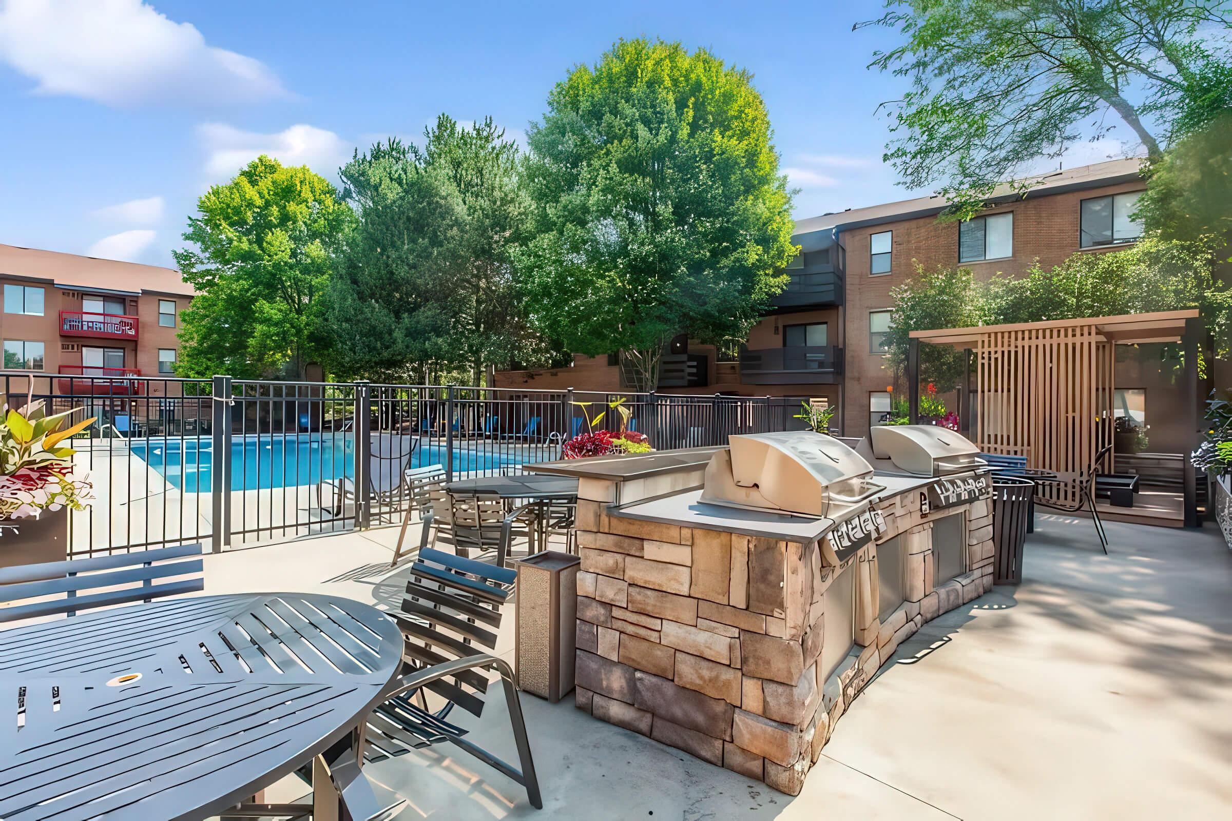 Outdoor pool area featuring a grill station, surrounded by stylish tables and chairs. Lush green trees provide shade, and a modern apartment building is visible in the background. The scene is bright and inviting, perfect for relaxation and gatherings.
