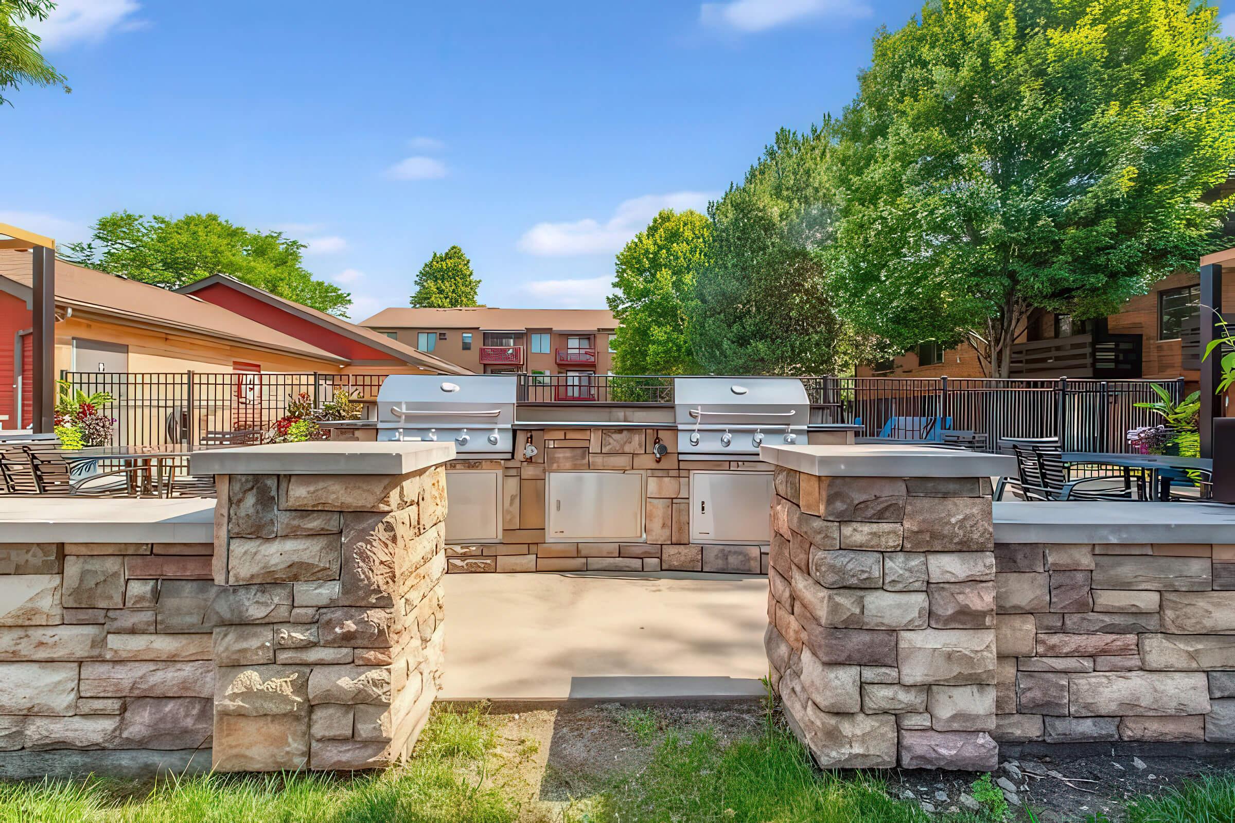 An outdoor barbecue area featuring two stainless steel grills surrounded by a stone wall. The space is adorned with green trees and shrubs, with a patio and outdoor seating visible in the background. Bright blue sky and well-maintained landscaping create a welcoming atmosphere for gatherings.