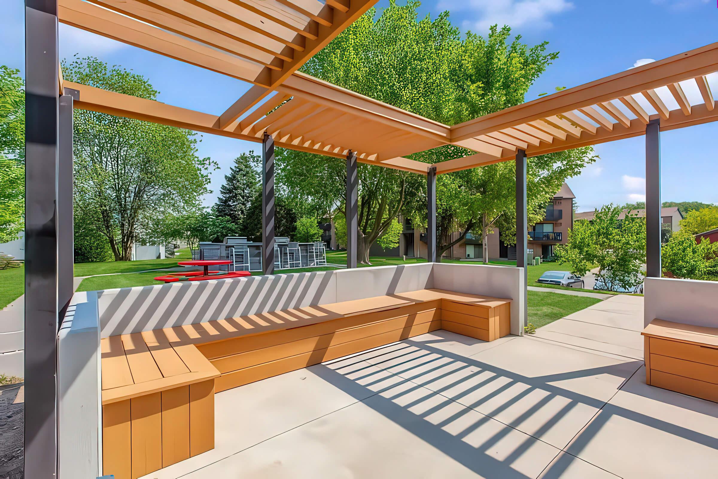 A modern outdoor seating area with orange wooden benches and a pergola, surrounded by green trees and grass. The space features a paved surface and is set against a backdrop of nearby buildings and park furniture. Bright blue sky fills the scene with soft, scattered clouds.