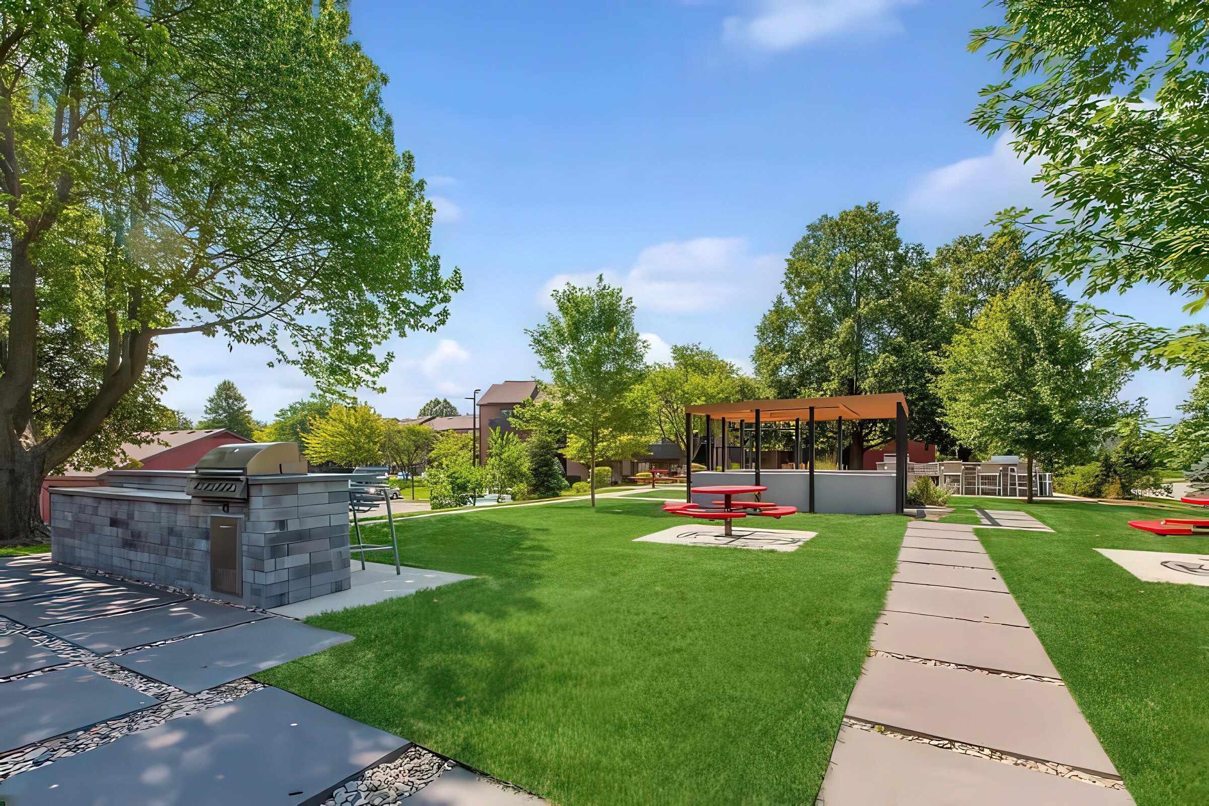 A green outdoor space featuring a grassy area with trees, picnic tables, and a covered pavilion. A stone grill is visible on one side, alongside a pathway made of flat stones leading through the park-like setting. The sky is clear with a few clouds, creating a bright and inviting atmosphere.