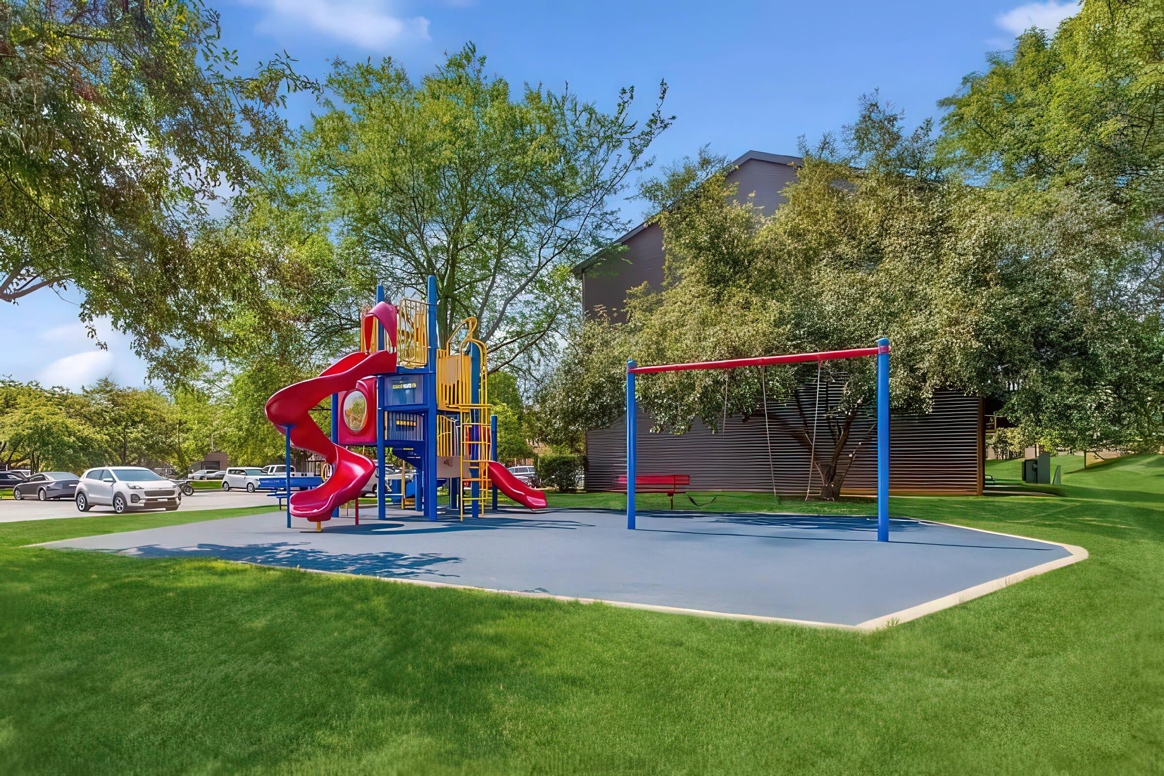 A colorful playground featuring a red slide and climbing structure, surrounded by green grass and trees. A swing set with two swings is also present. In the background, cars are parked along a pathway, and a building can be seen partially obscured by trees under a clear blue sky.