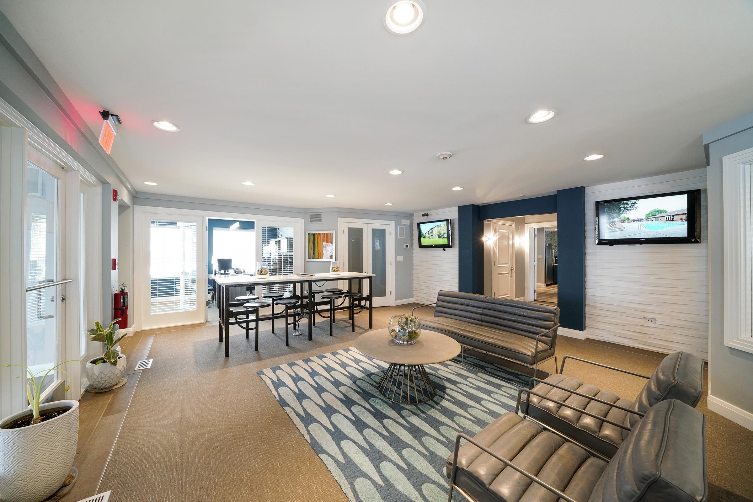 A modern lounge area featuring a light-colored interior with a patterned rug, comfortable seating, and a coffee table. There are two wall-mounted televisions and large windows allowing natural light. In the background, a kitchenette with bar seating is visible.