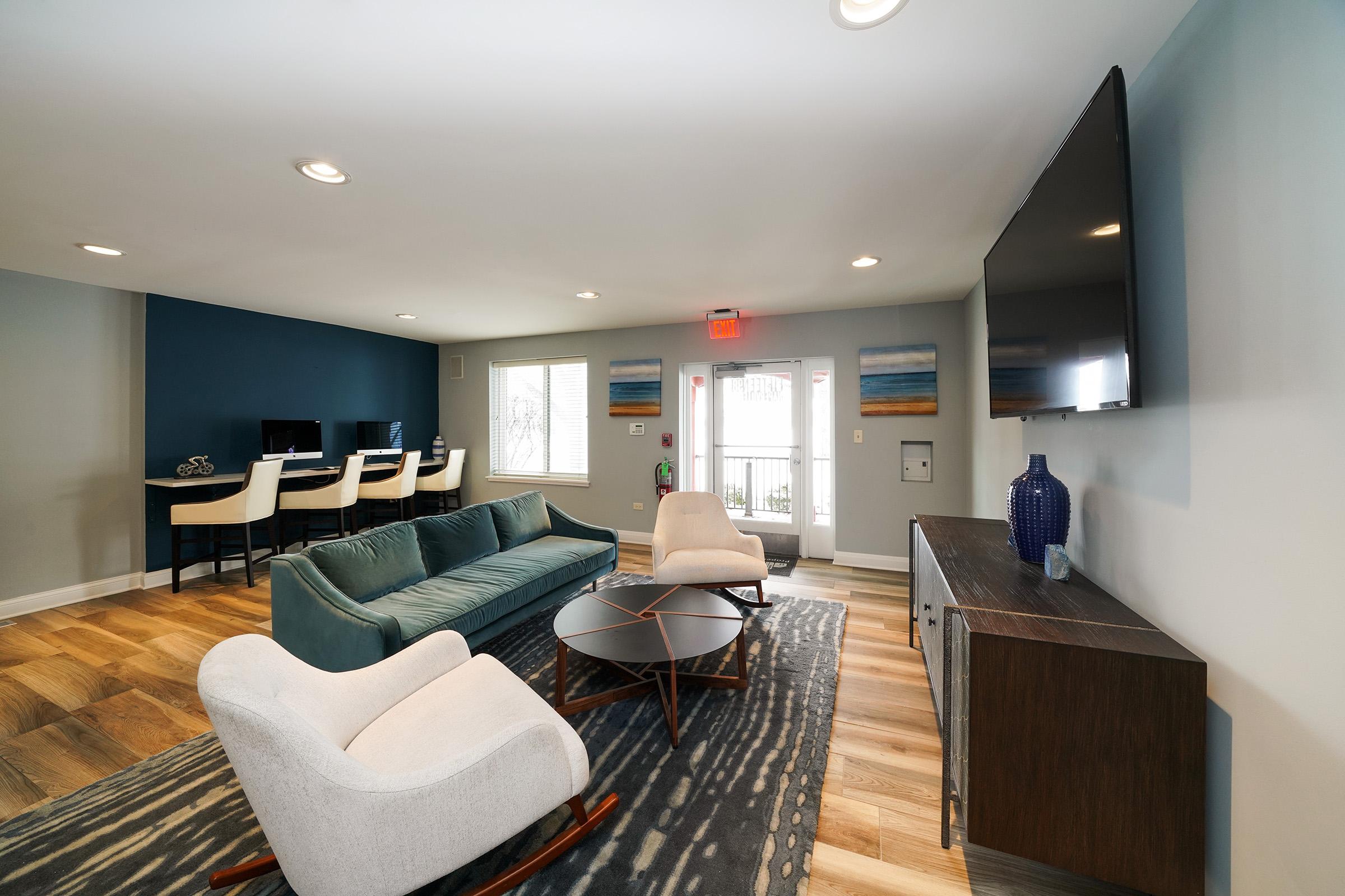 Modern living room with a comfortable teal sofa, a stylish round coffee table, and two cozy armchairs. The space features hardwood flooring, a flat-screen TV mounted on the wall, and a window with natural light. A dining area with high chairs is visible in the background, creating a welcoming atmosphere.