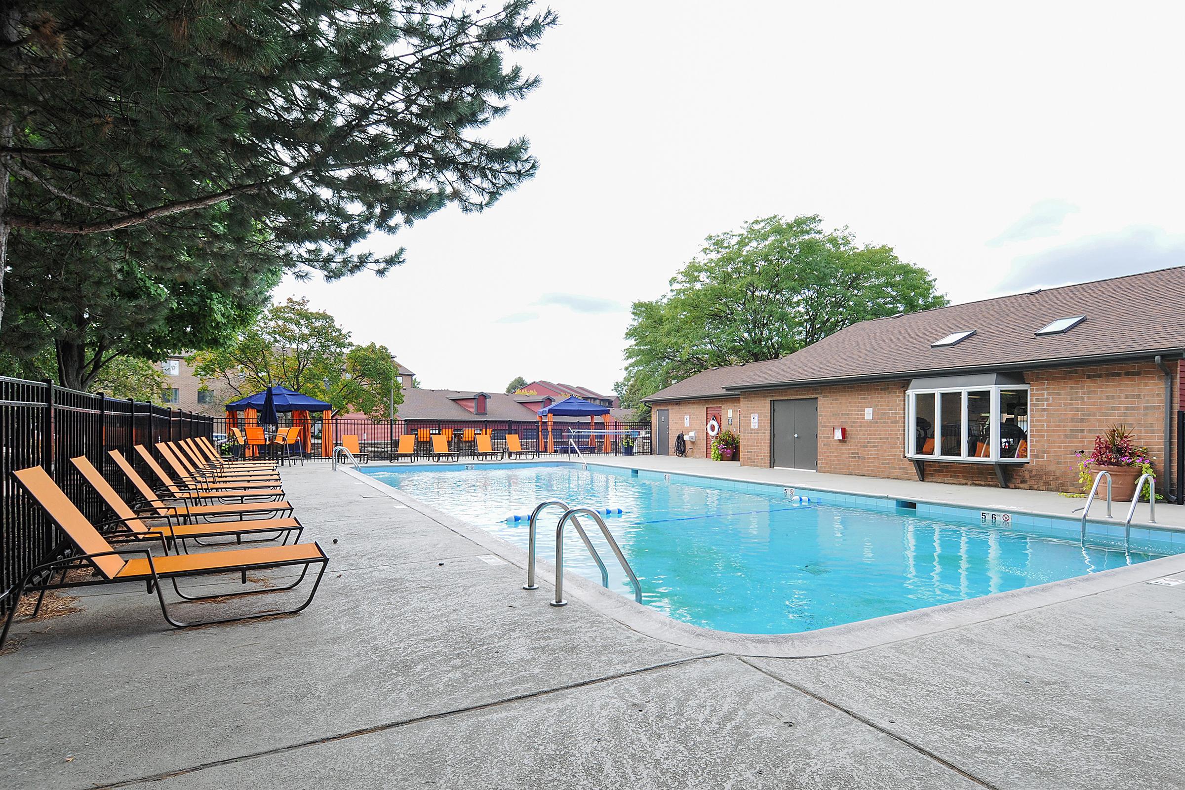 A well-maintained outdoor swimming pool surrounded by lounge chairs. The pool area features a fence, sun loungers positioned along the deck, and shaded seating under blue umbrellas. A brick building with windows is visible in the background, along with trees and landscaping. The sky is partly cloudy.