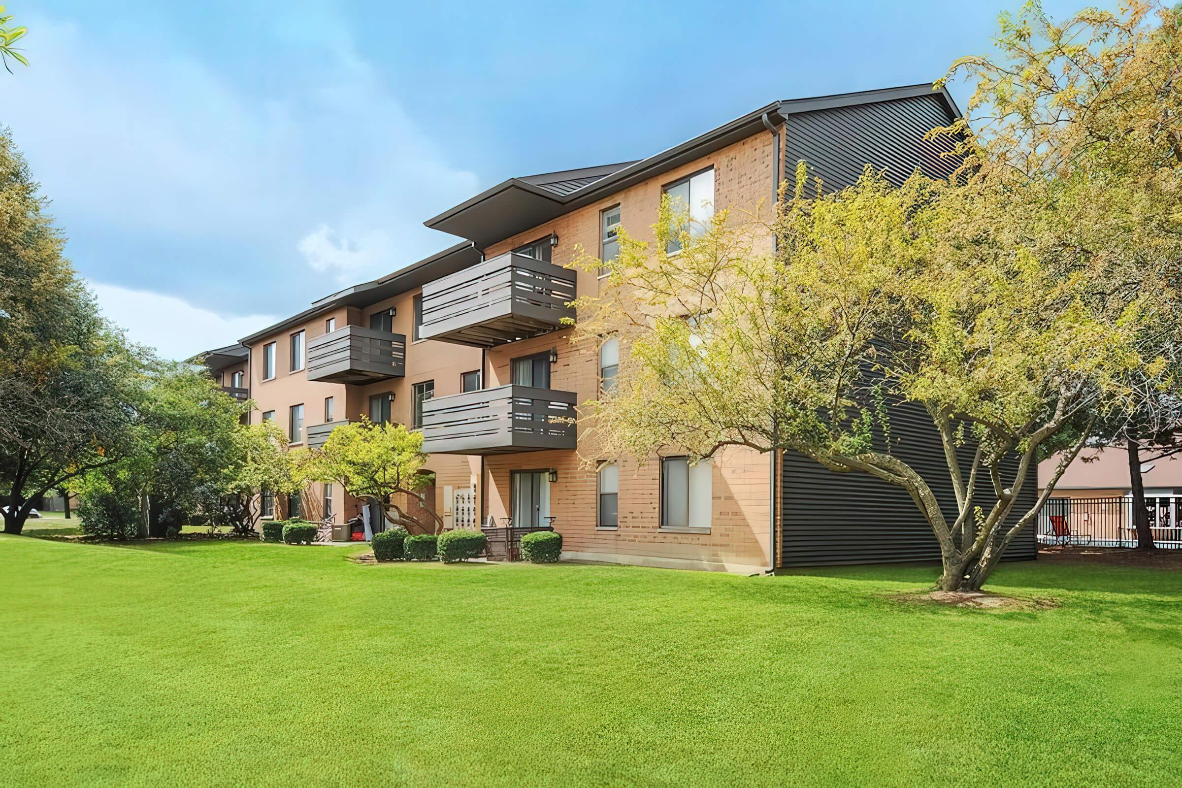 A multi-story apartment building with a brick exterior and balconies, surrounded by well-maintained green lawns and trees. The sky is partly cloudy, creating a pleasant atmosphere in the residential area.