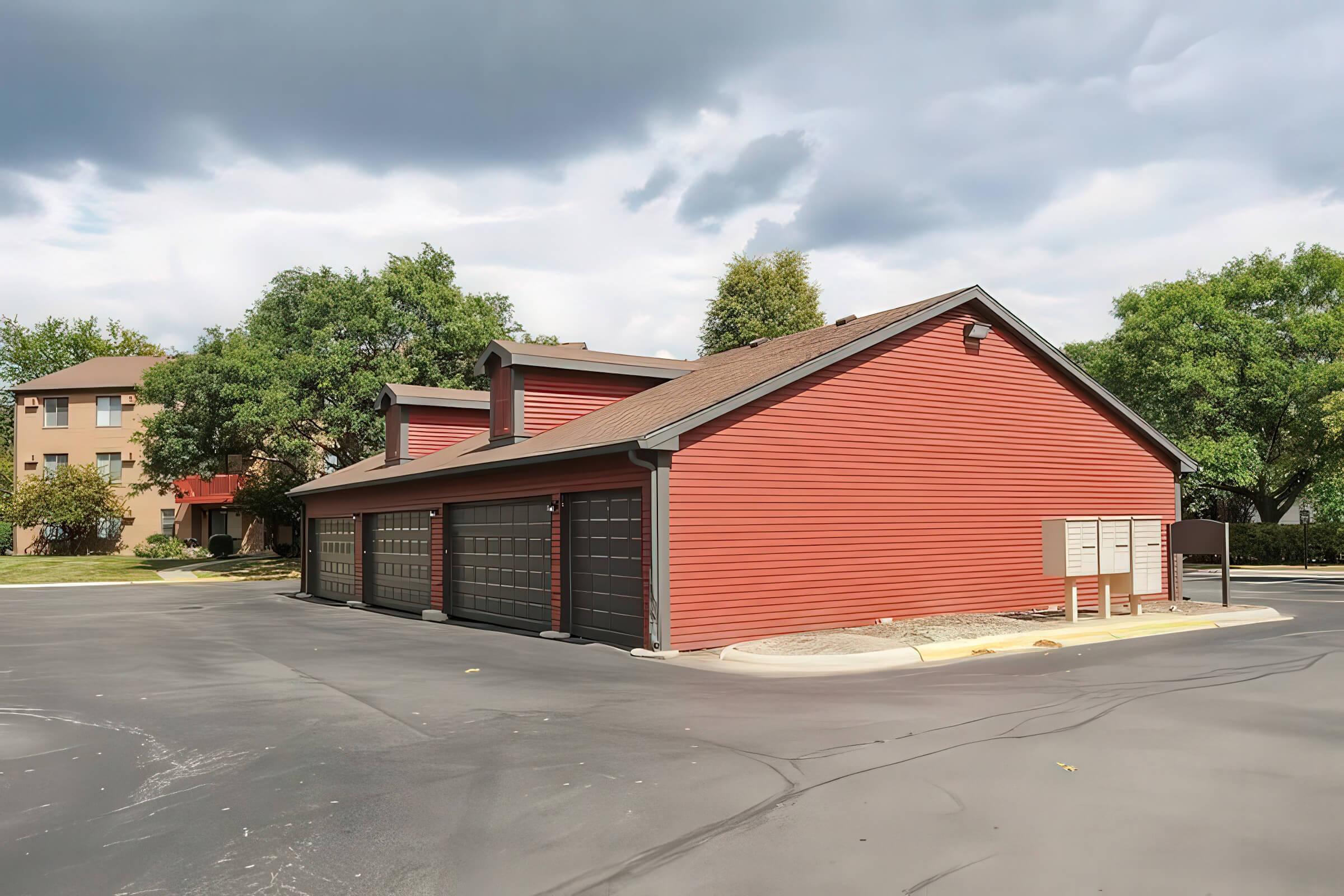 A red single-story building with a sloped roof and multiple garage doors, situated in a paved parking area. Nearby, there are trees and additional residential buildings in the background under a cloudy sky.