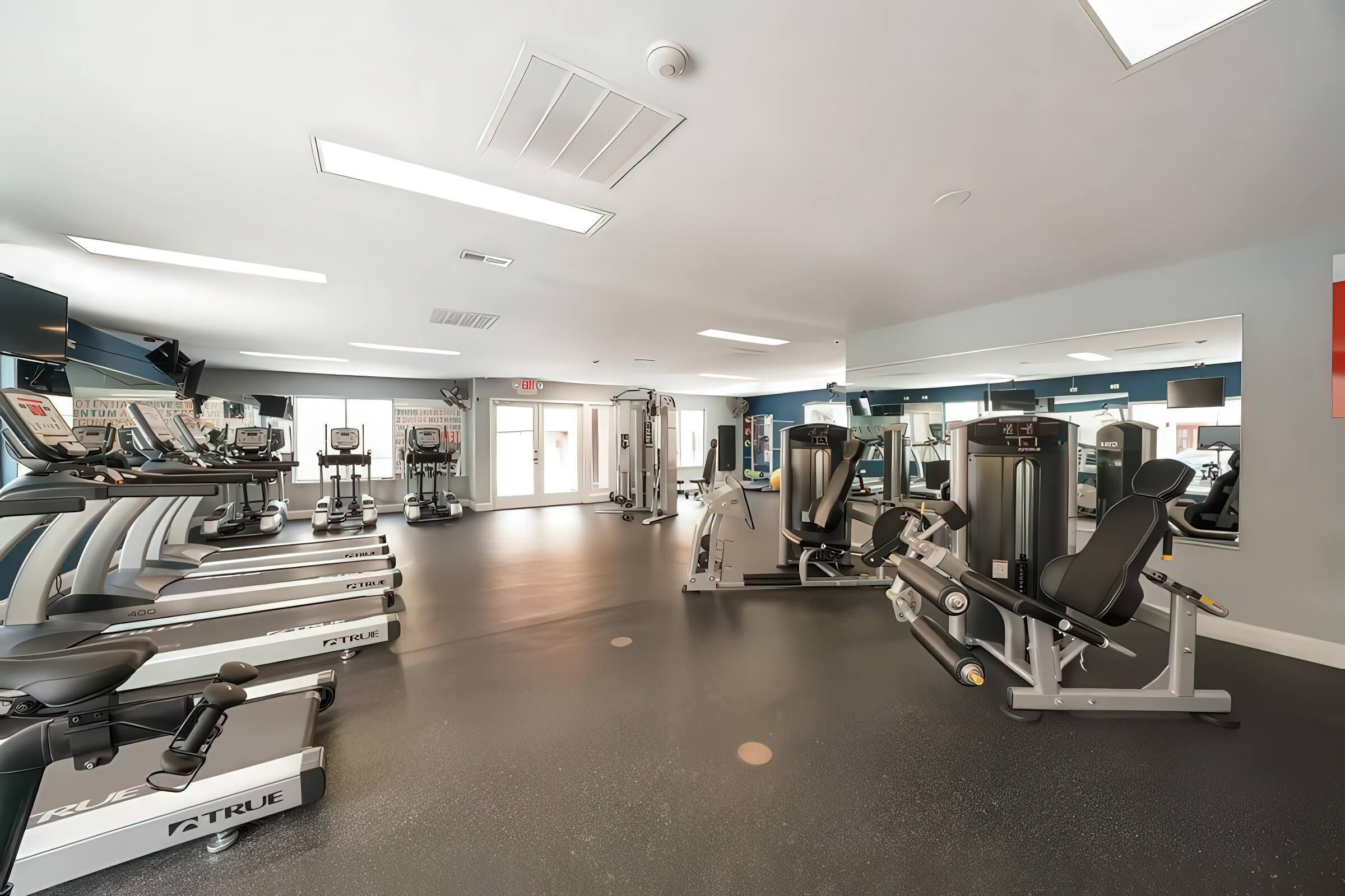 A spacious fitness room featuring various exercise equipment including treadmills, stationary bikes, and weight machines. Large mirrors reflect the equipment, and there is ample natural light entering through windows. The floor is made of a black rubber material for safety and comfort.