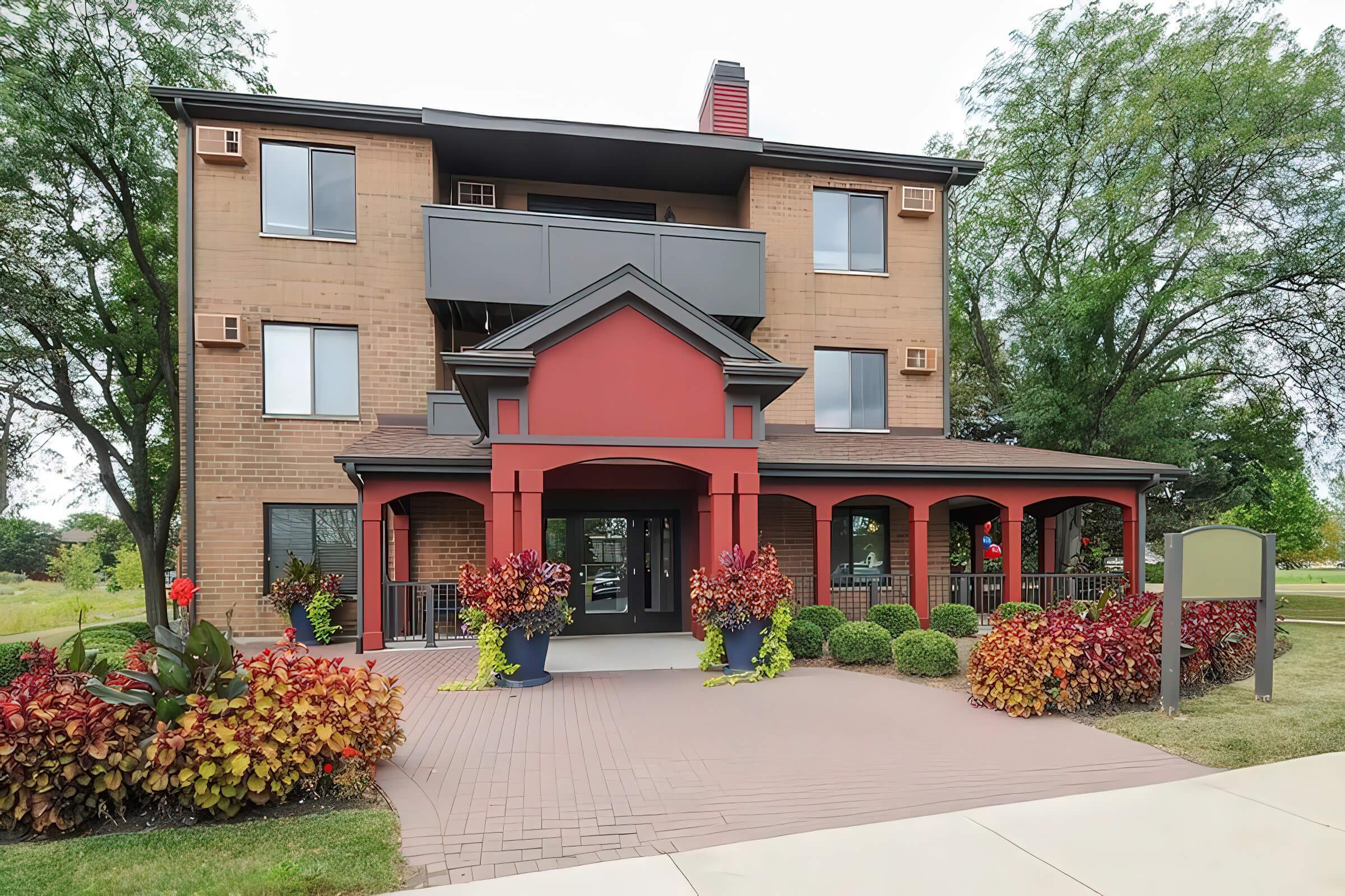 A three-story building featuring a red entrance archway, large windows, and a covered porch with decorative potted plants. Surrounding the building are colorful flower beds and well-maintained greenery, creating a welcoming atmosphere. The sky is partly cloudy.