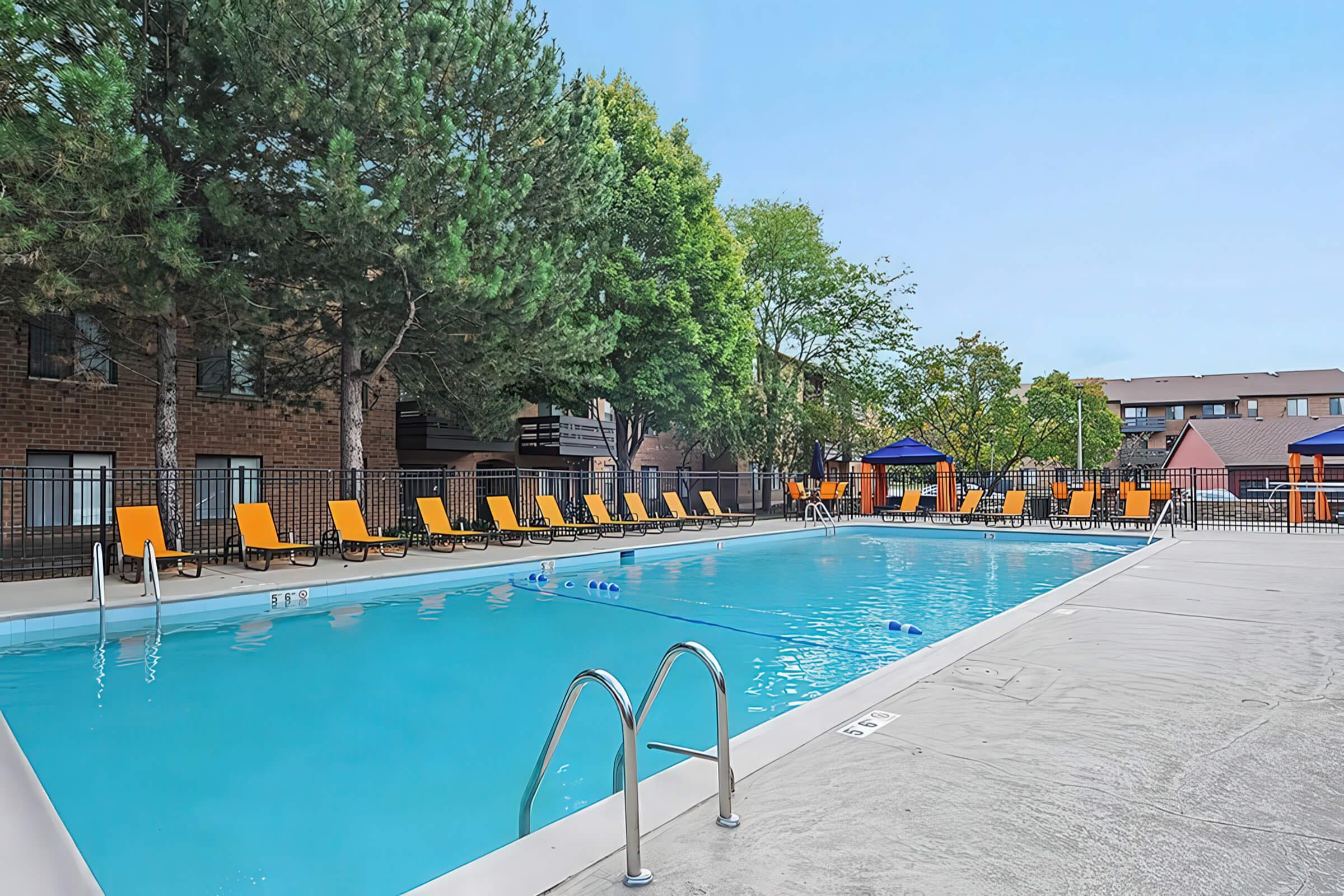 A spacious outdoor swimming pool surrounded by lounge chairs and trees. Several blue floats are in the water. The pool area features shaded cabanas and is located near apartment buildings, creating a relaxing atmosphere for residents and visitors. Clear blue sky overhead.