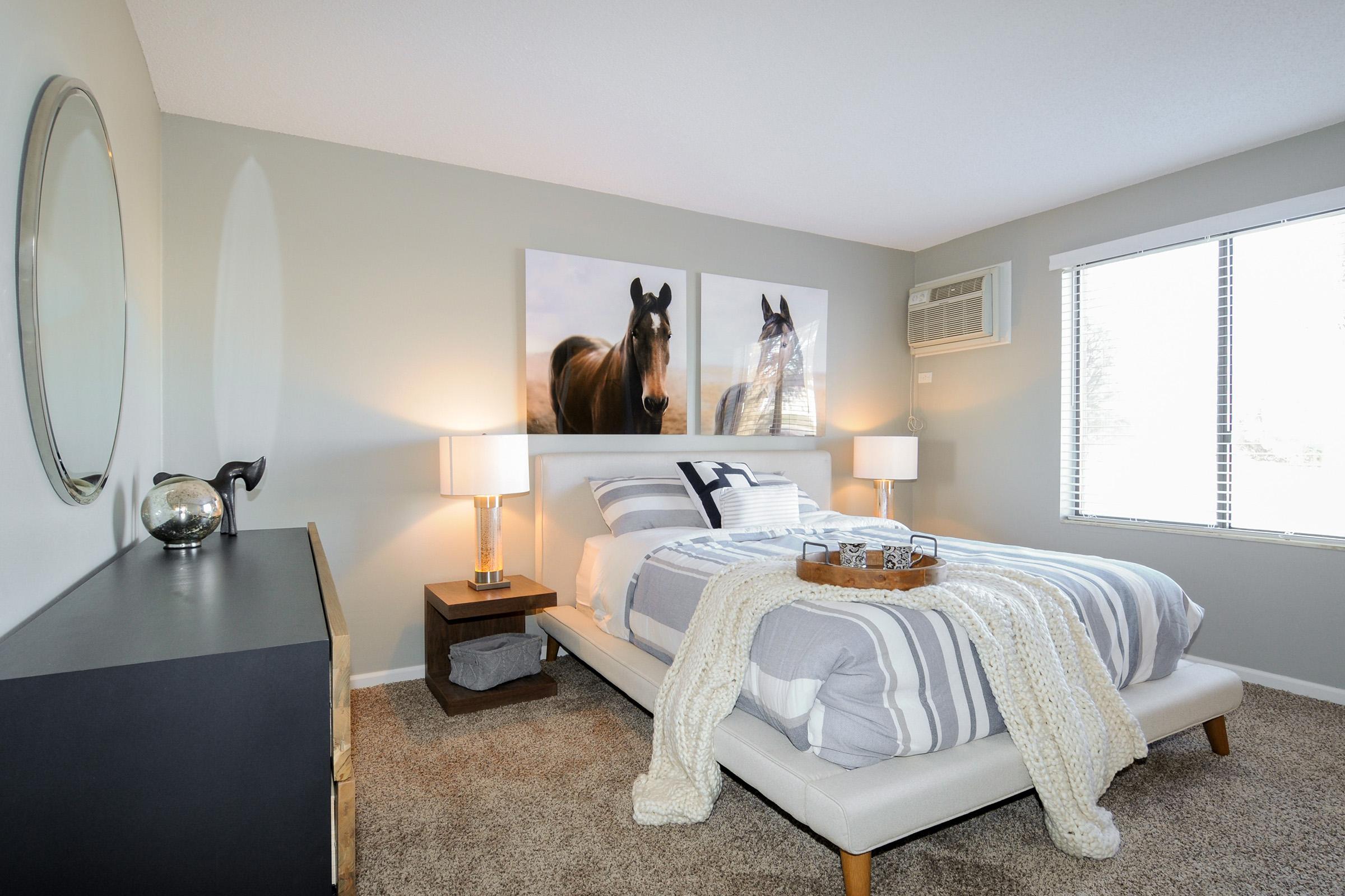A cozy bedroom featuring a light gray color scheme, a neatly made bed with gray and white striped bedding, two framed horse prints on the wall, an air conditioning unit, a round mirror, and two bedside lamps on either side of a dark wood nightstand with a tray.