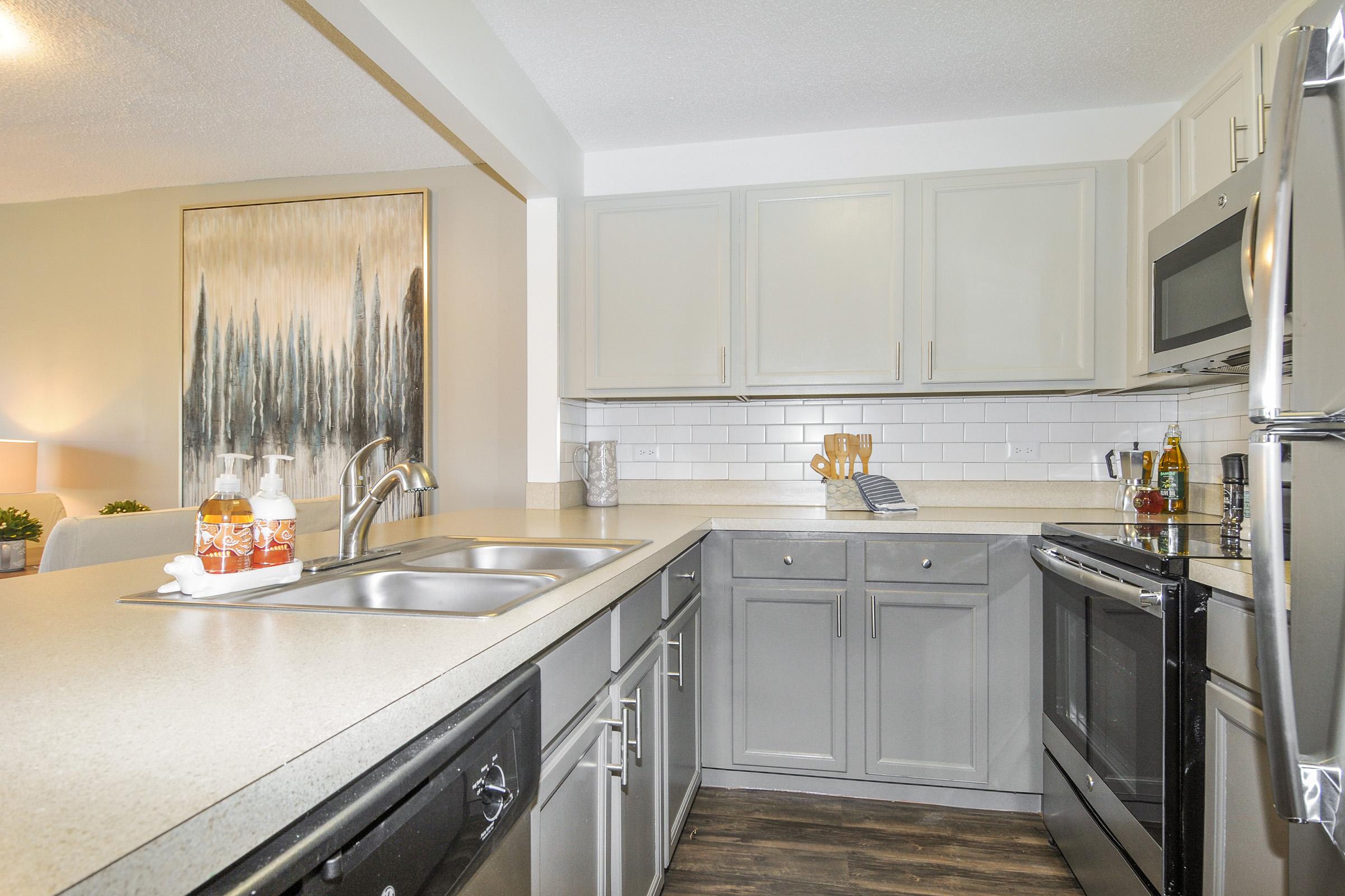 A modern kitchen featuring gray cabinetry, a stainless steel microwave, and a double sink. The countertop is light-colored with dish soap and a towel neatly arranged. A framed artwork depicting trees is hanging on the wall, and there are plants and accessories on the countertop, enhancing the warm ambiance.