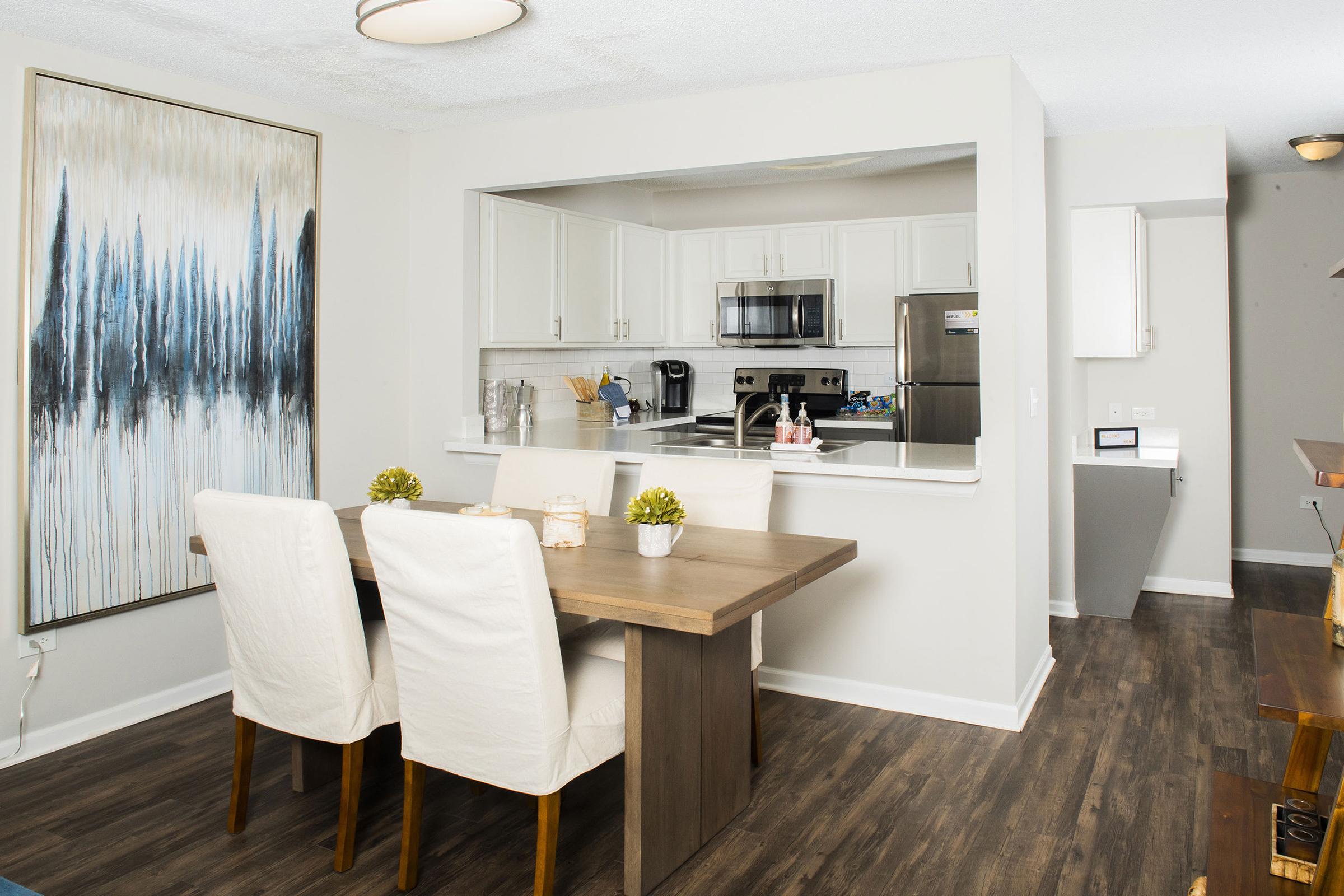 A modern kitchen and dining area featuring a wooden table with two white chairs, a large abstract painting on the wall, and stainless steel appliances visible in the kitchen. The flooring is dark wood, and the space is well-lit with natural and artificial light.