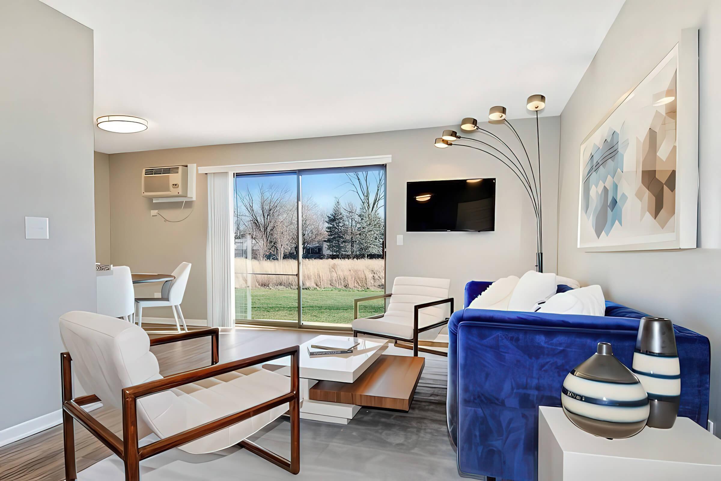 A modern living room featuring a blue sofa, white chairs, and a wooden coffee table. A large sliding glass door offers a view of a grassy area outside. An air conditioning unit is mounted on the wall, and a television is mounted above a side table. Decorative items include a vase and artwork on the wall.