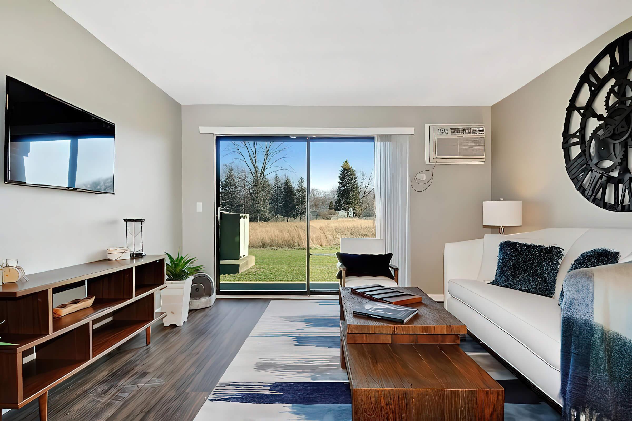 A cozy living room featuring a white sofa with blue accents, a wooden coffee table, and a dark wooden TV stand. A large window with sliding doors offers a view of a grassy outdoor area. A flat-screen TV is mounted on the wall, and there is a decorative wall clock nearby, enhancing the modern decor.