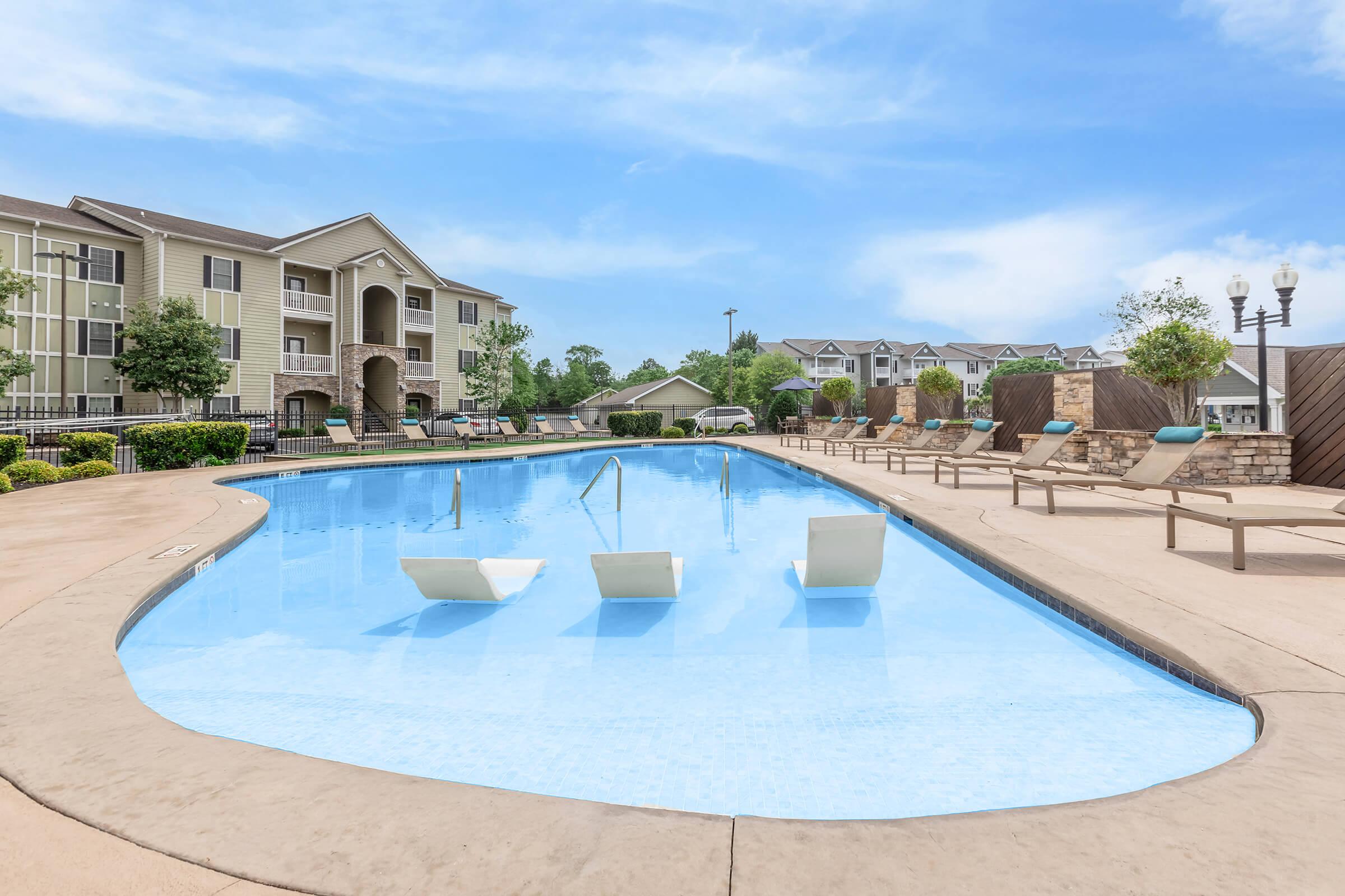 A swimming pool with clear blue water surrounded by lounge chairs and landscaped greenery. In the background, there are beige apartment buildings. A sunny sky is visible overhead, creating a bright and inviting atmosphere.