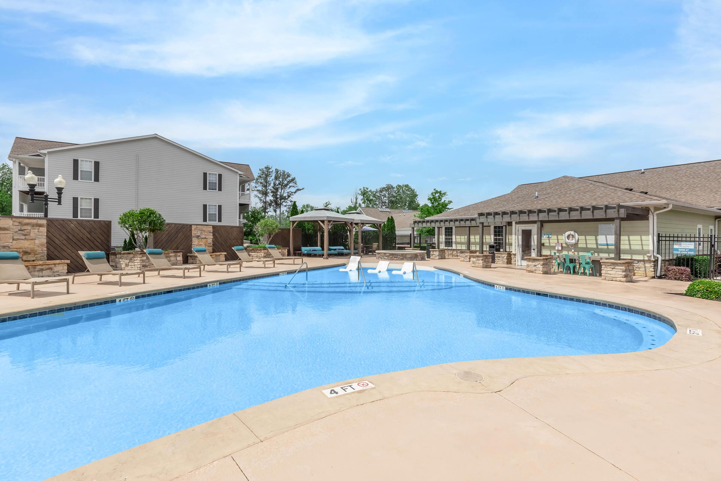 A clear blue swimming pool surrounded by lounge chairs and cabanas. The pool area features light-colored concrete decking, with a backdrop of residential buildings and green landscaping under a bright blue sky.