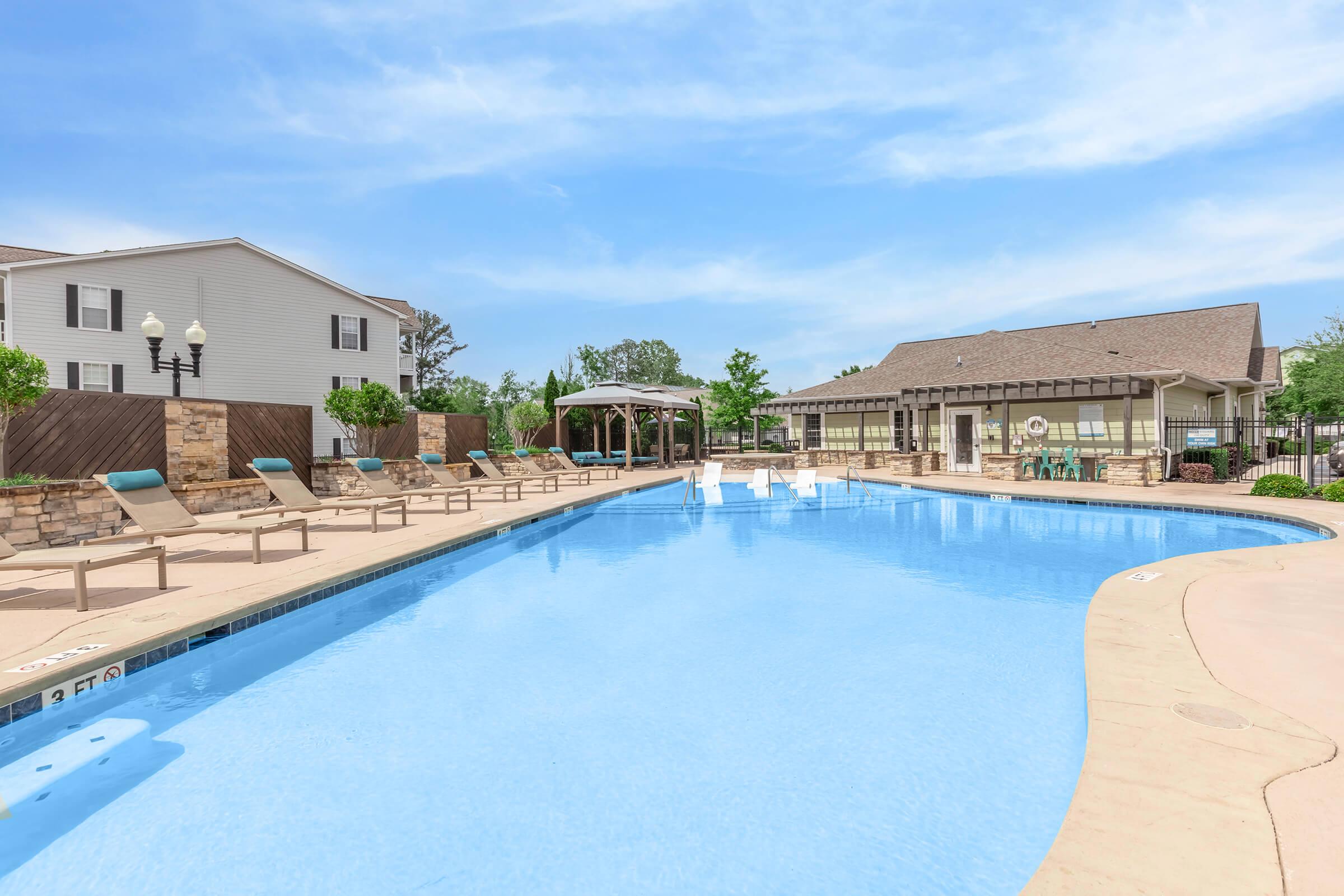 A clear blue swimming pool surrounded by lounge chairs, with a gazebo and landscaped areas nearby. The scene includes residential buildings in the background under a bright sky.