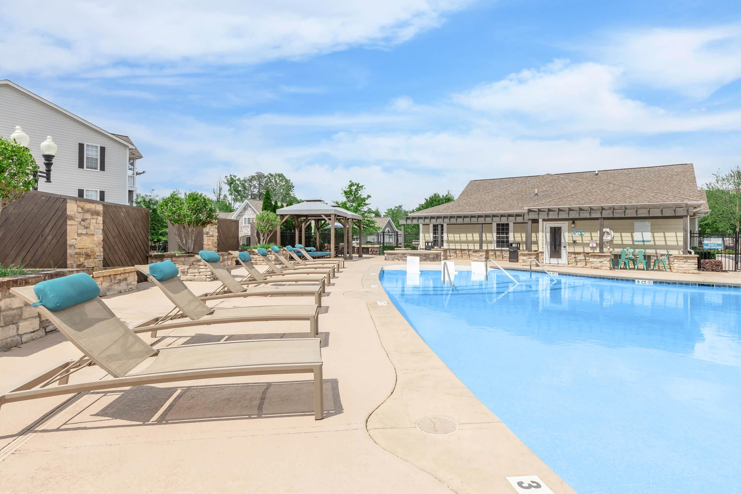 A serene outdoor swimming pool area with lounge chairs arranged neatly along the edge. The pool is crystal clear, surrounded by trees and buildings, with a shaded gazebo in the background. The sky is bright blue with a few fluffy clouds, creating a relaxing atmosphere.