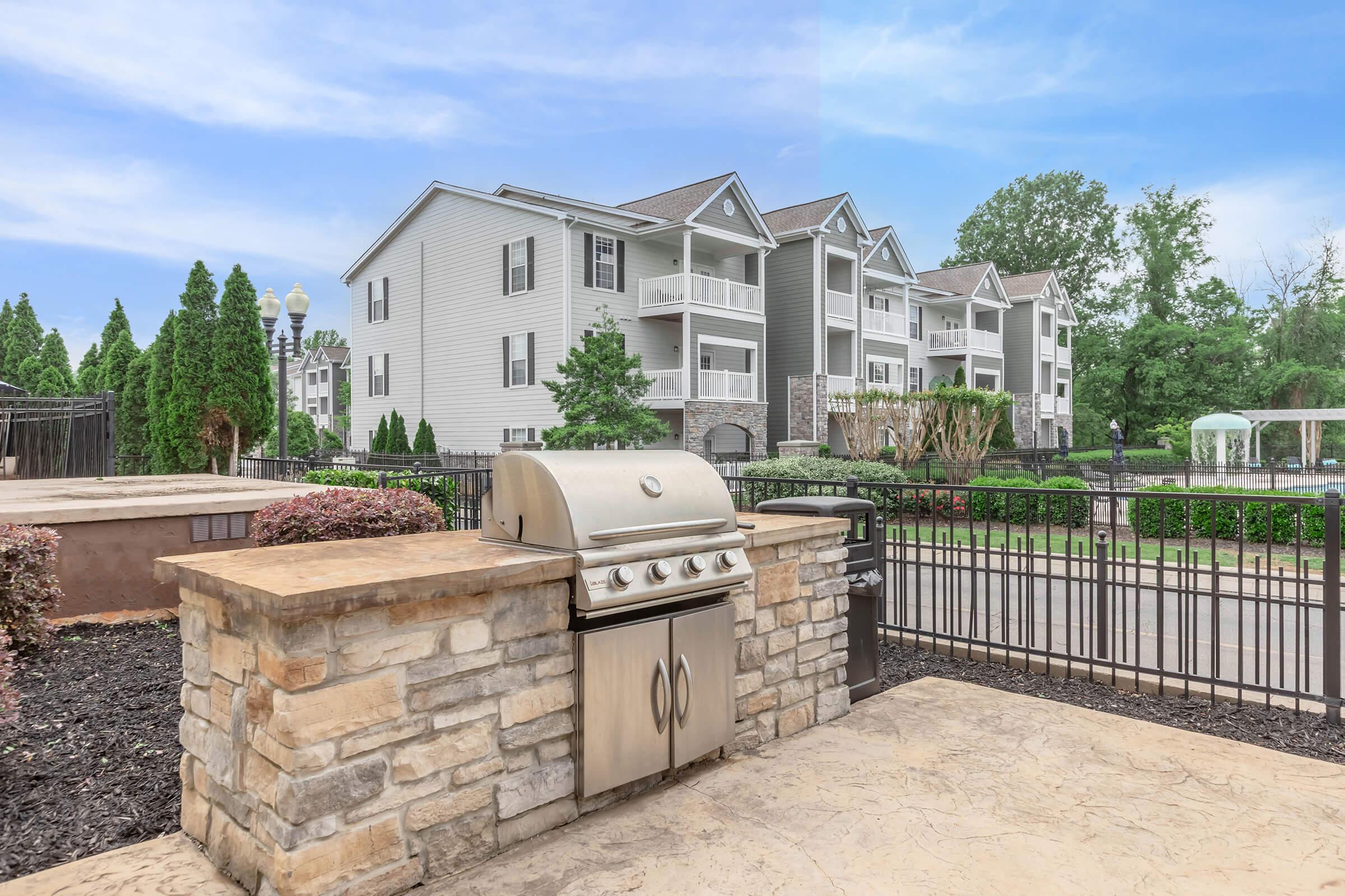 A stainless steel grill on a stone patio in front of a modern apartment building. The building features multiple balconies and is surrounded by landscaped greenery. A gated area and blue sky complete the setting, suggesting a recreational space for residents.