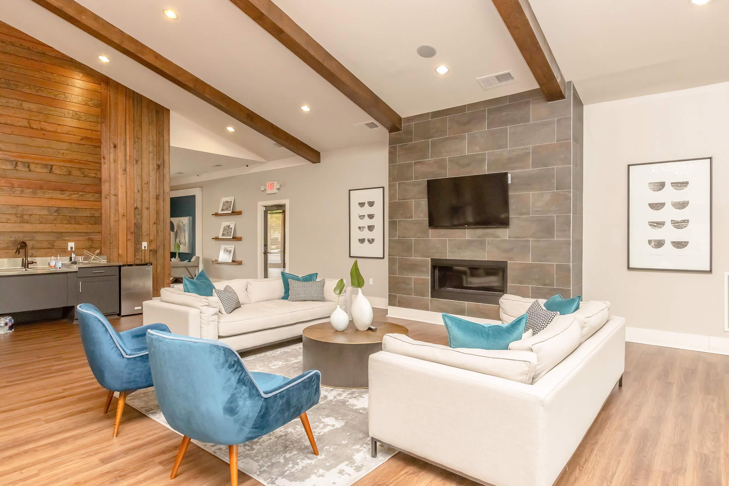 A modern living room featuring two light-colored sofas with teal accent pillows, a round coffee table, and two blue accent chairs. The room has wooden beams on the ceiling, a stone fireplace, a wall-mounted TV, and art prints on display. A kitchen area is visible in the background.