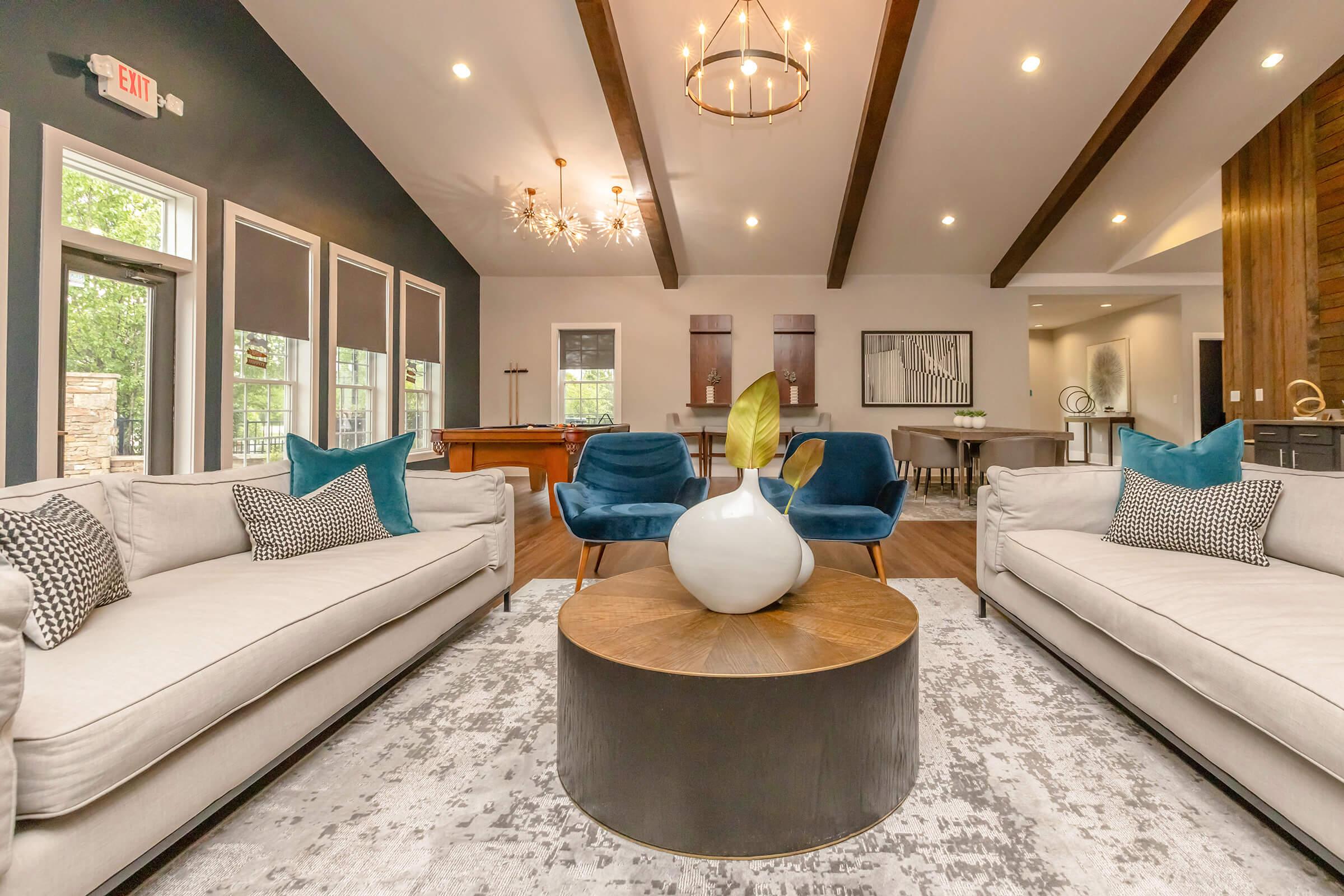 A modern living room featuring two light-colored sofas with decorative cushions, a round wooden coffee table, and two blue accent chairs. The room has high ceilings with wooden beams, stylish light fixtures, and large windows letting in natural light, creating a cozy and inviting atmosphere.