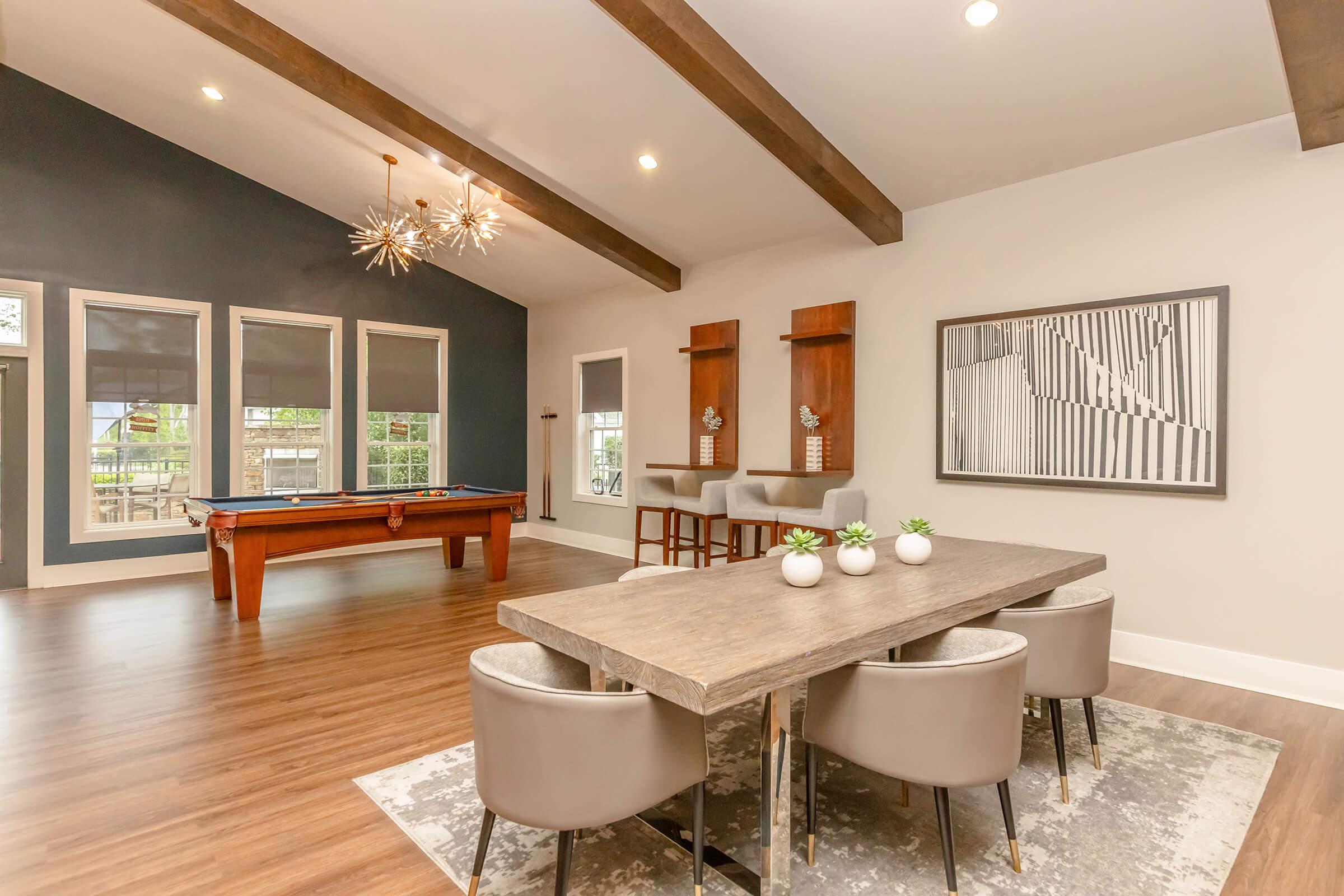 A modern, stylish interior featuring a dining area with a wooden table and gray upholstered chairs. In the background, there is a pool table and large windows. The ceiling has exposed beams, and decorative light fixtures add elegance. A geometric art piece is displayed on the wall.