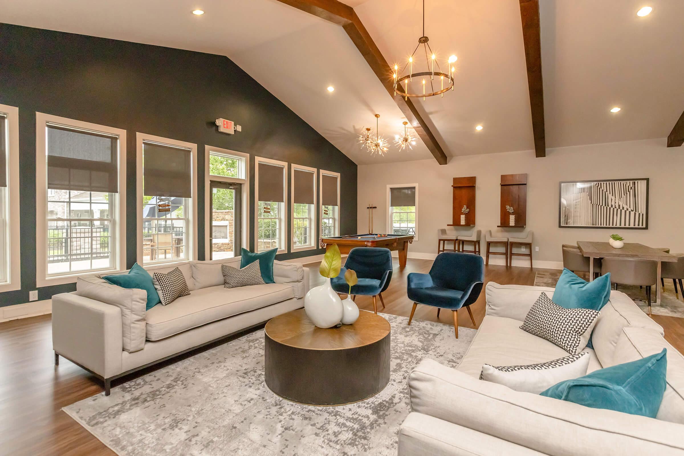 A stylish living room featuring two plush sofas with blue and white accents, a round coffee table, and modern armchairs. The space has high ceilings with wooden beams, large windows letting in natural light, and contemporary decor, creating a warm and inviting atmosphere.
