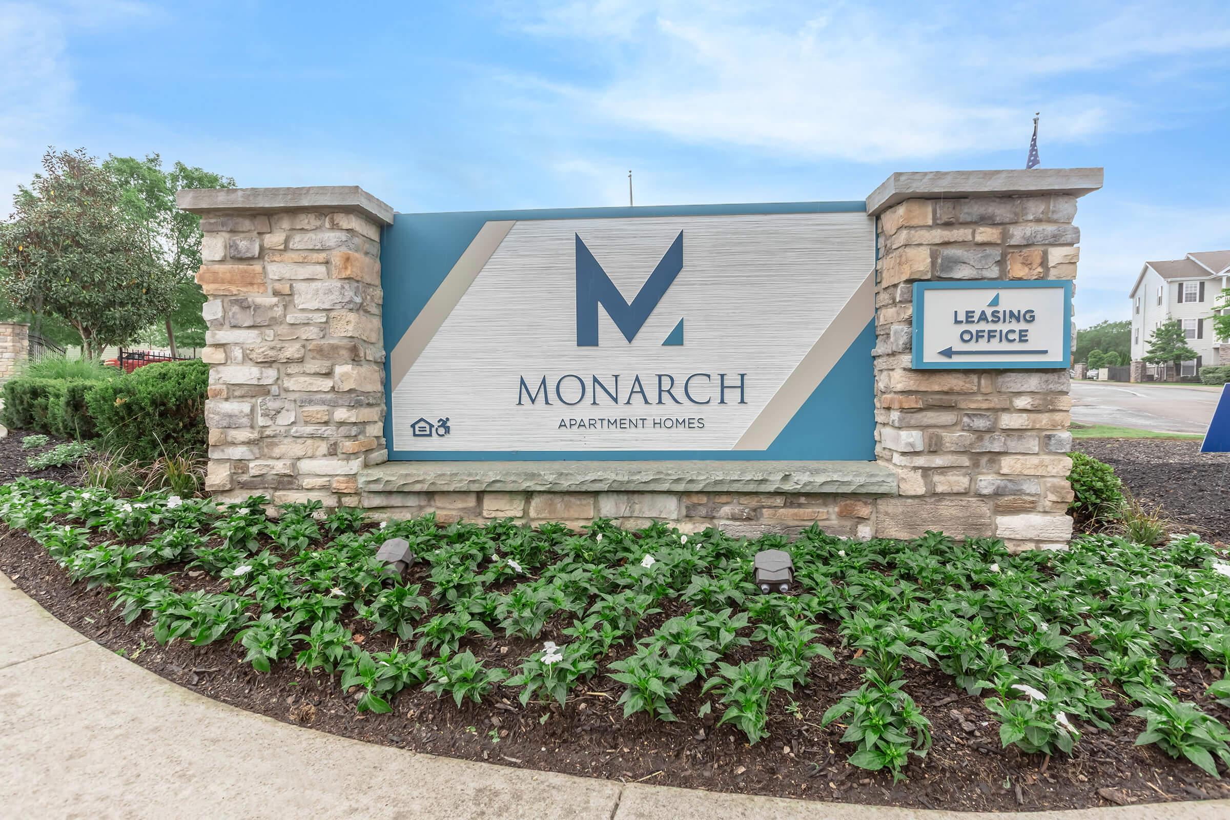 Sign for Monarch Apartment Homes featuring the name "MONARCH" prominently displayed in a modern font, surrounded by a stone structure and landscaped with green plants. A smaller sign on the right indicates "Leasing Office."