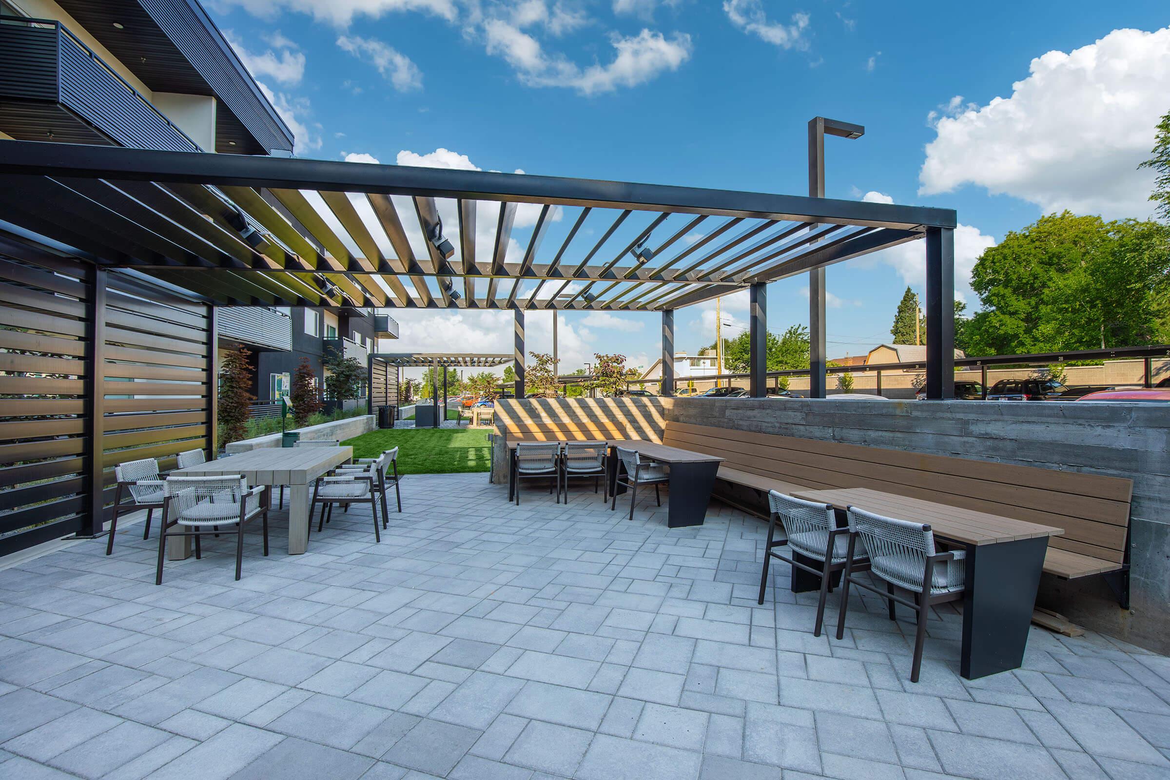 A modern outdoor patio area with a pergola overhead, featuring light-colored stone flooring. There are several tables and chairs arranged for dining, along with a bench along one side. Lush greenery and trees are visible in the background under a bright blue sky with scattered clouds.