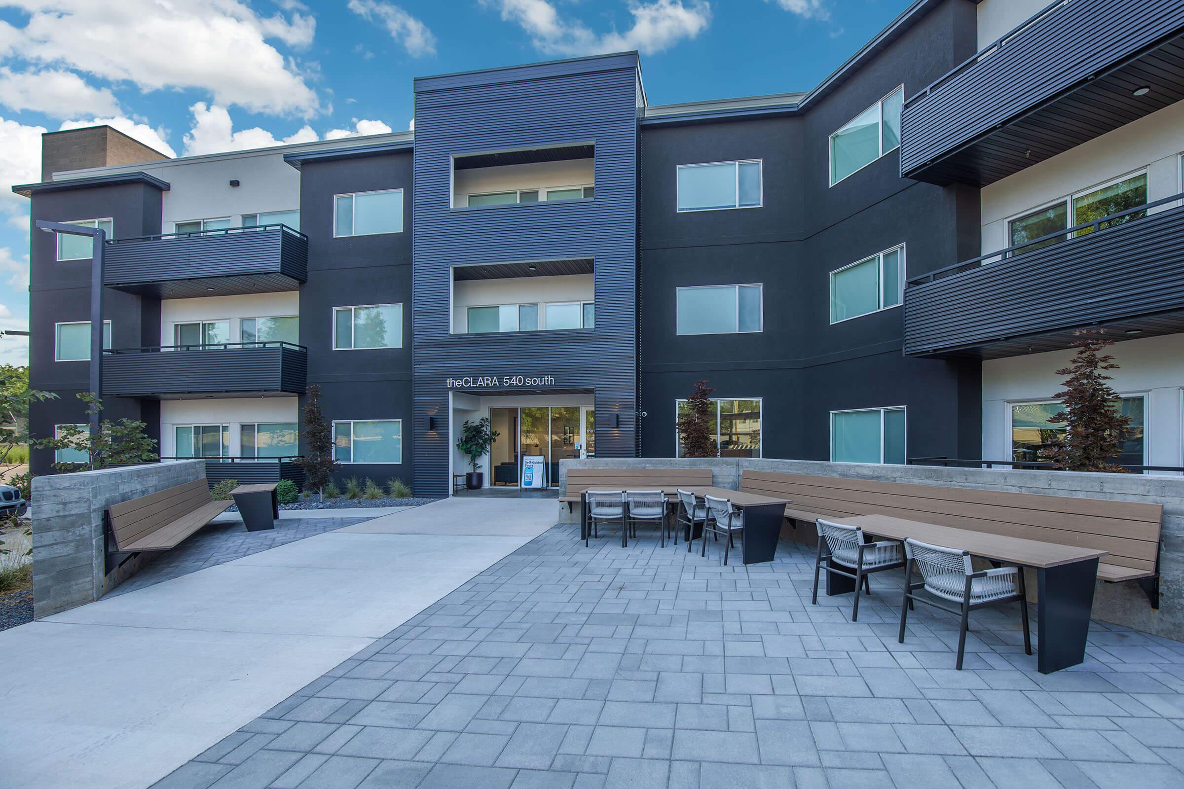 Modern apartment building with multiple balconies and large windows. A paved pathway leads to the entrance, which features a sign that reads "theOLARA 549 south." On the left side, there are outdoor seating areas with wooden benches and chairs, surrounded by greenery under a partly cloudy sky.