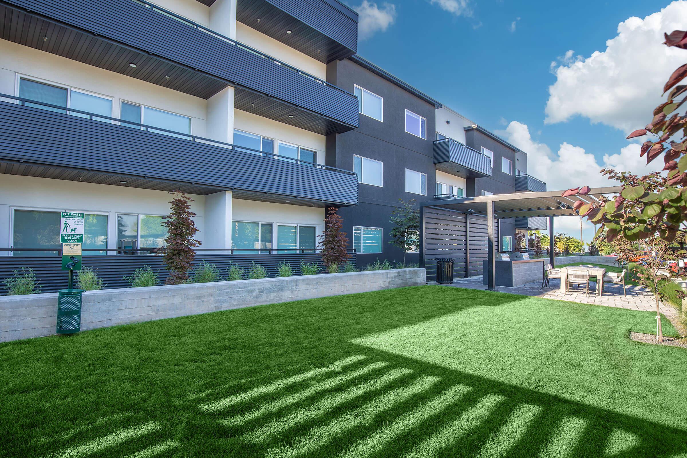 Modern apartment building with a sleek design, featuring multiple balconies and large windows. In the foreground, a well-maintained green lawn with landscaped plants. To the side, there is a shaded patio area with seating, surrounded by trees and a clear blue sky with fluffy clouds.